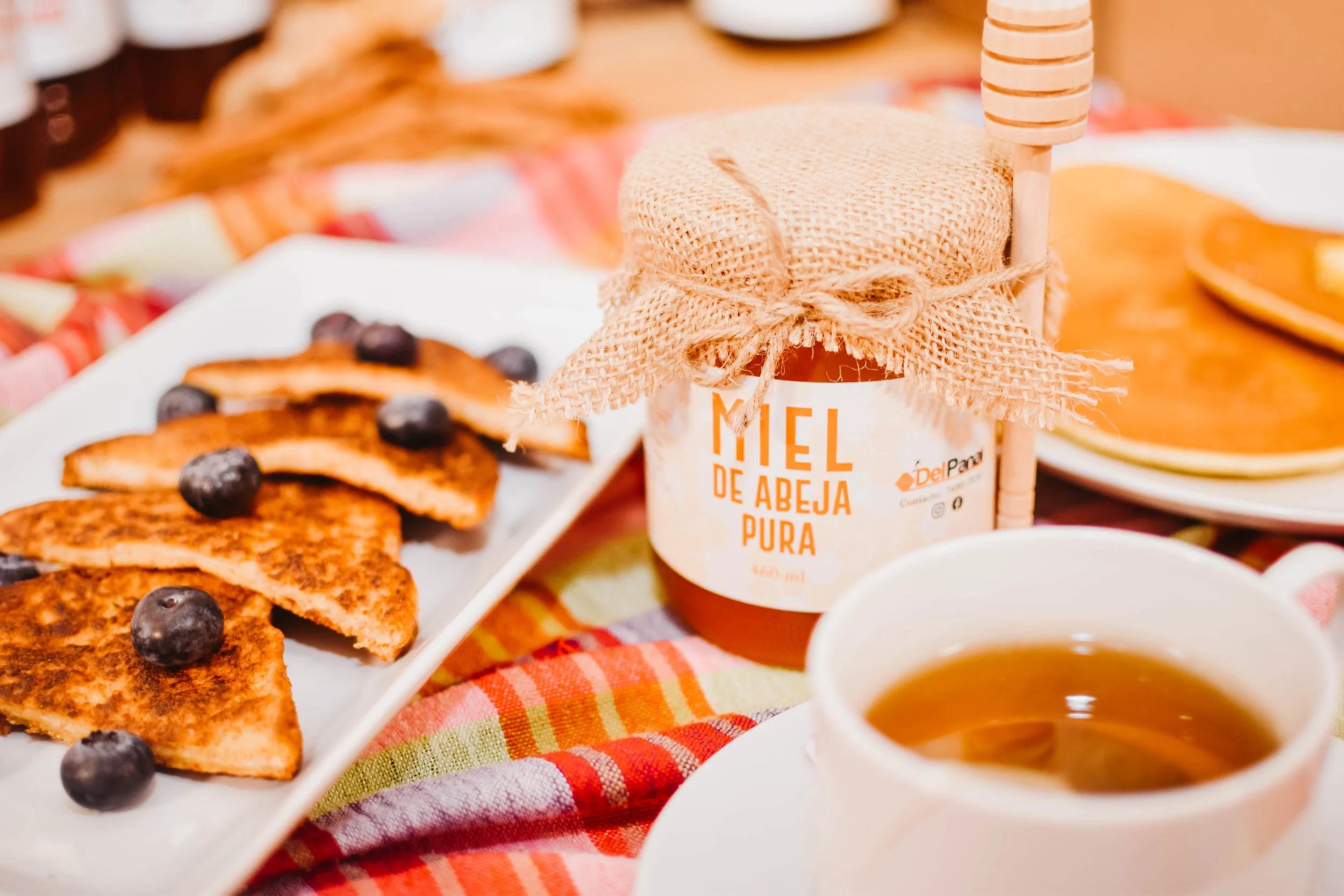 Miel de abeja pura en frasco con tapa de tela, acompañado de galletas con arándanos y tazas de té o café en un ambiente acogedor.