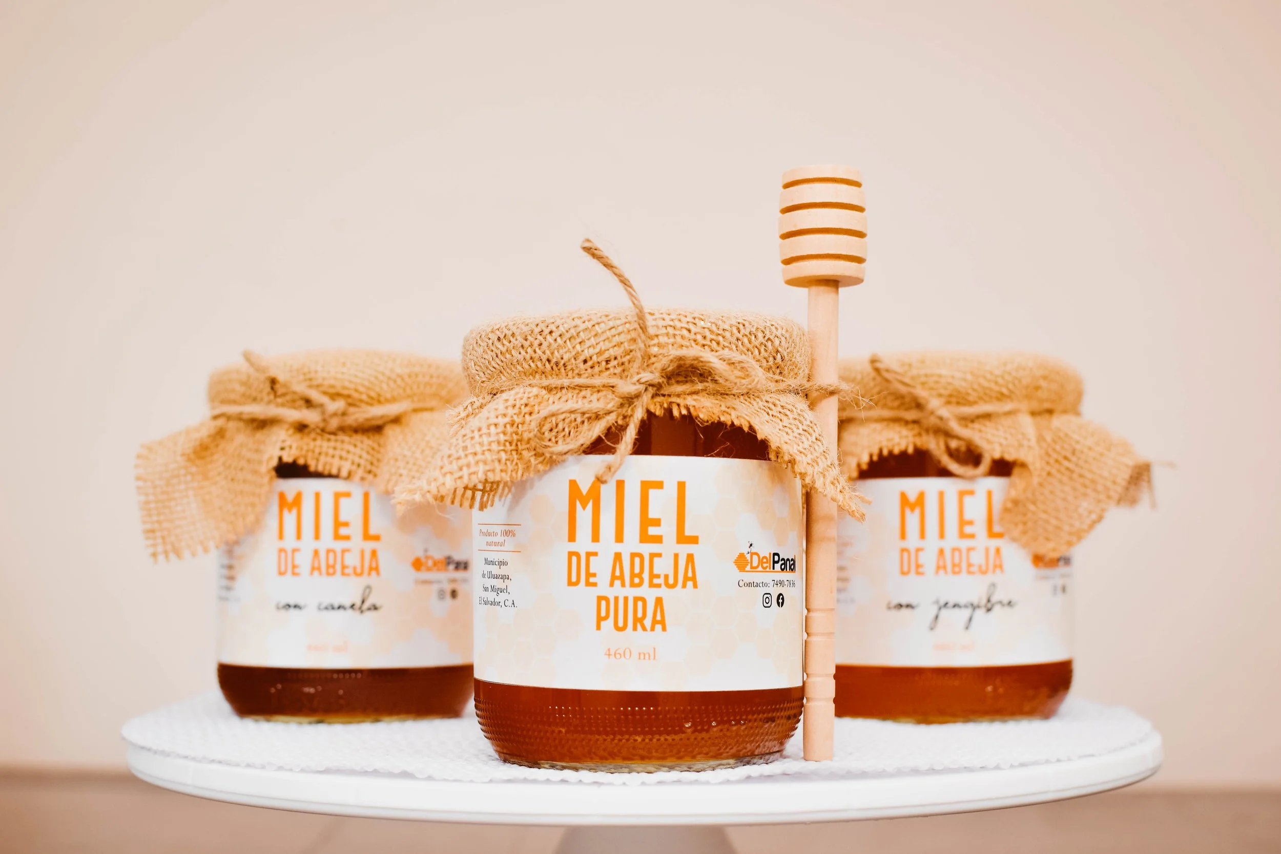 Tres frascos de miel de abeja con etiquetas en español, decorados con telas de arpillera y cinta, acompañados de un palo de madera para miel, sobre una base blanca.
