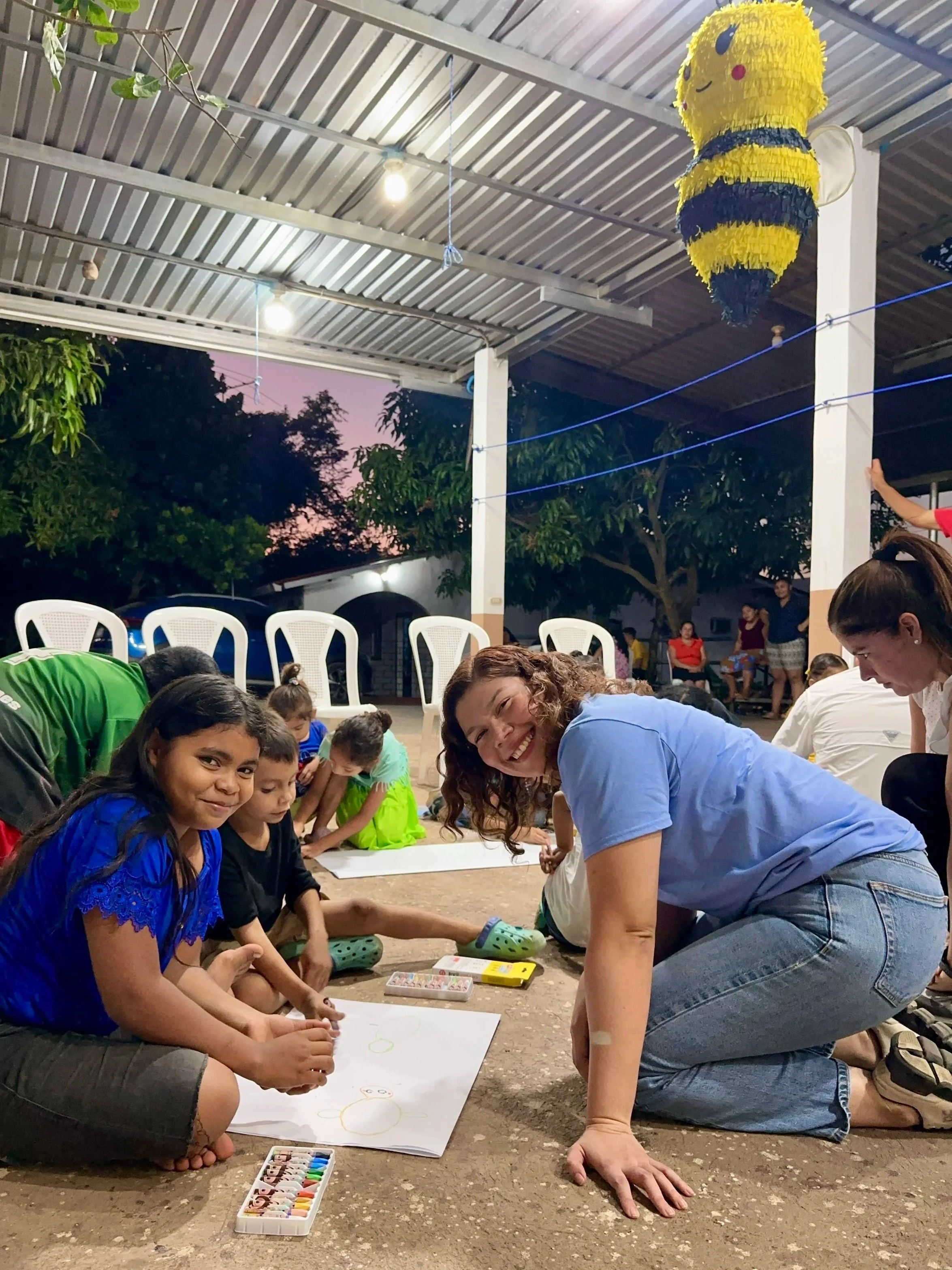 Niños y una mujer participando en una actividad de dibujo en un espacio abierto cubierto, con decoraciones y personas observando en el fondo.