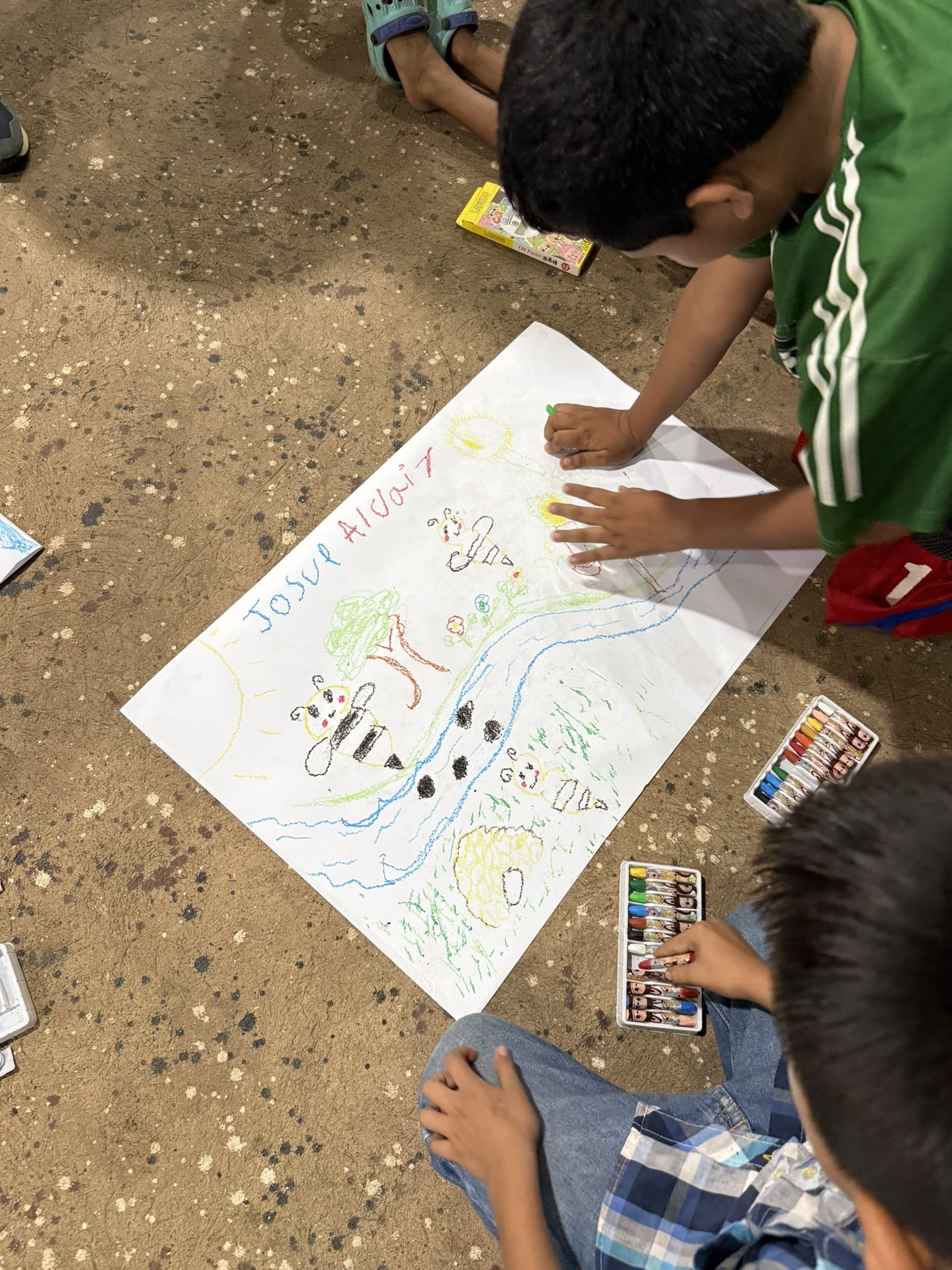 Niños creando un dibujo en una hoja grande, rodeados de crayones y otros niños observando.