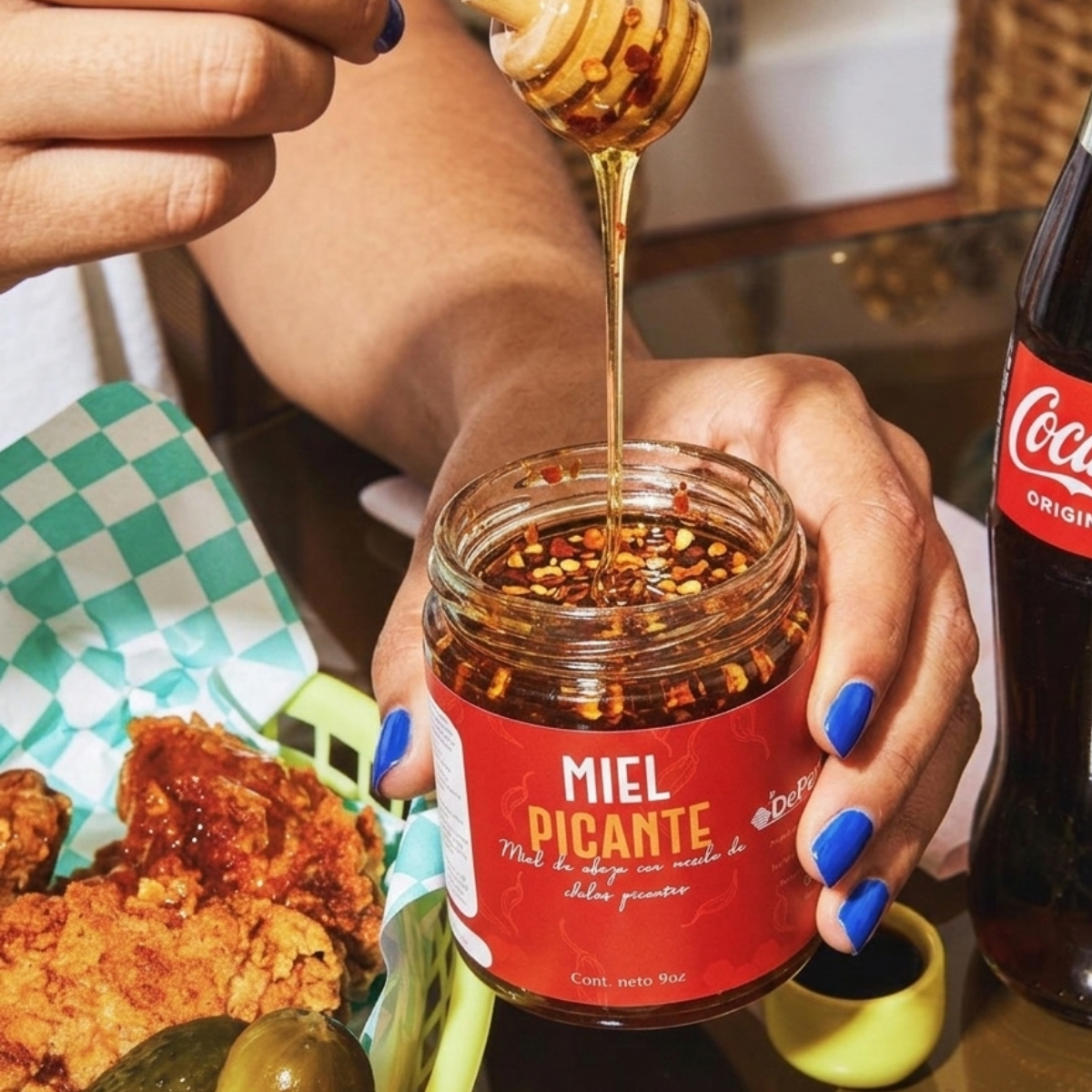 Una mano sostiene un frasco de miel picante, con miel mostrándose al ser vertida en el frasco. Al fondo hay pollo frito, un pepino y un vaso de Coca-Cola.