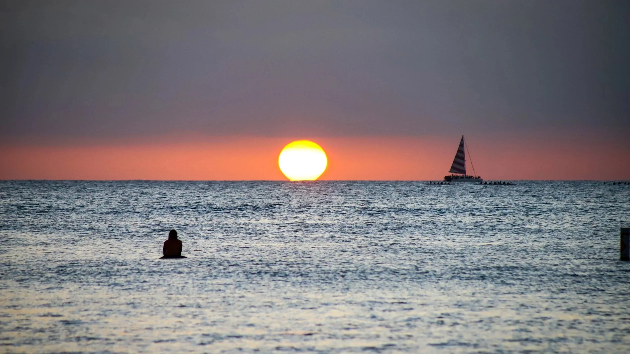 A person sitting at the beach and looking at the sunset with a sailboat in the distance.