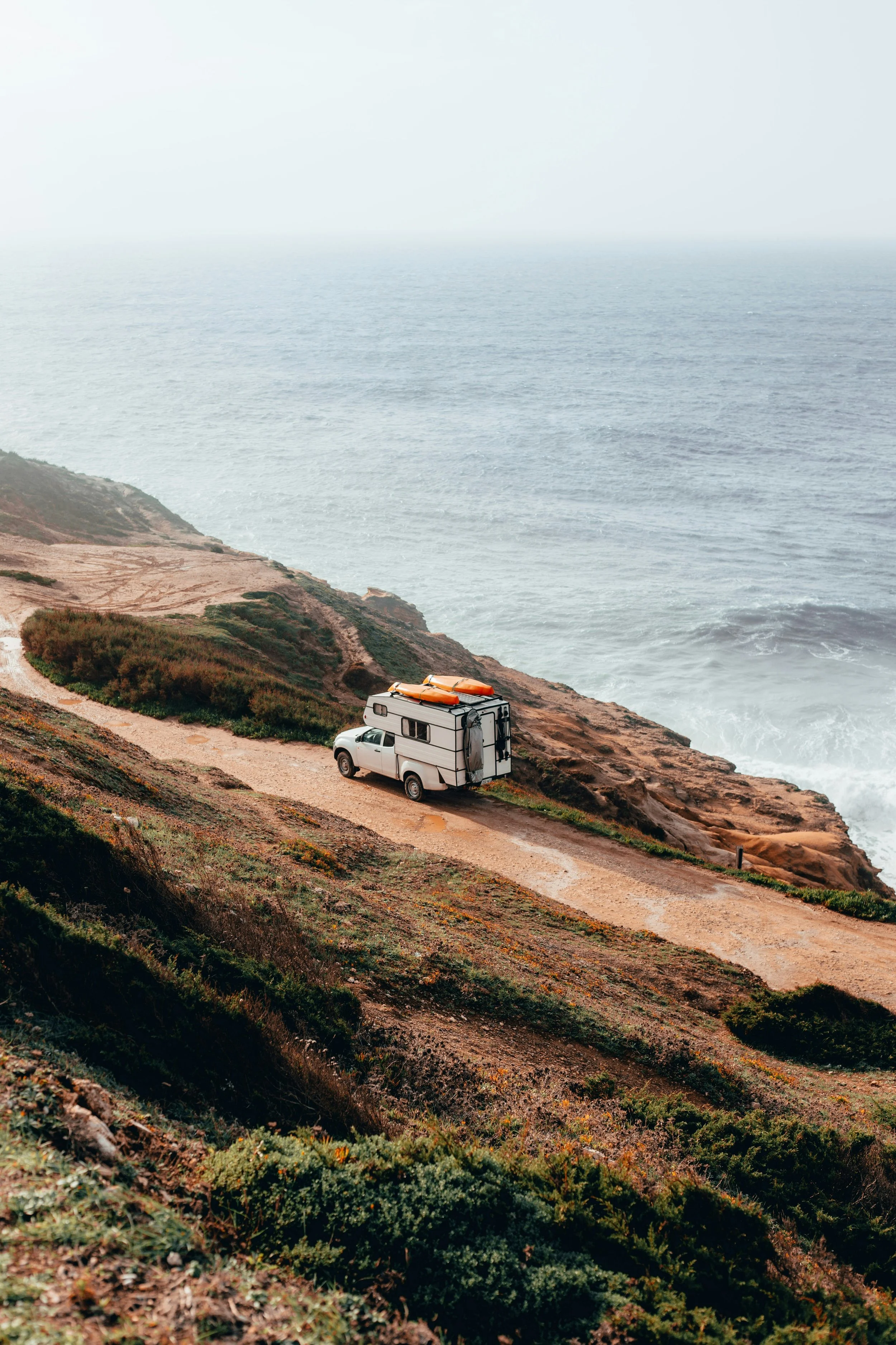 Un vehículo recreativo estacionado en un camino de tierra junto a un acantilado que mira al océano, con vegetación baja en primer plano y cielo nublado en el fondo. mueble camper camper thevanbox
