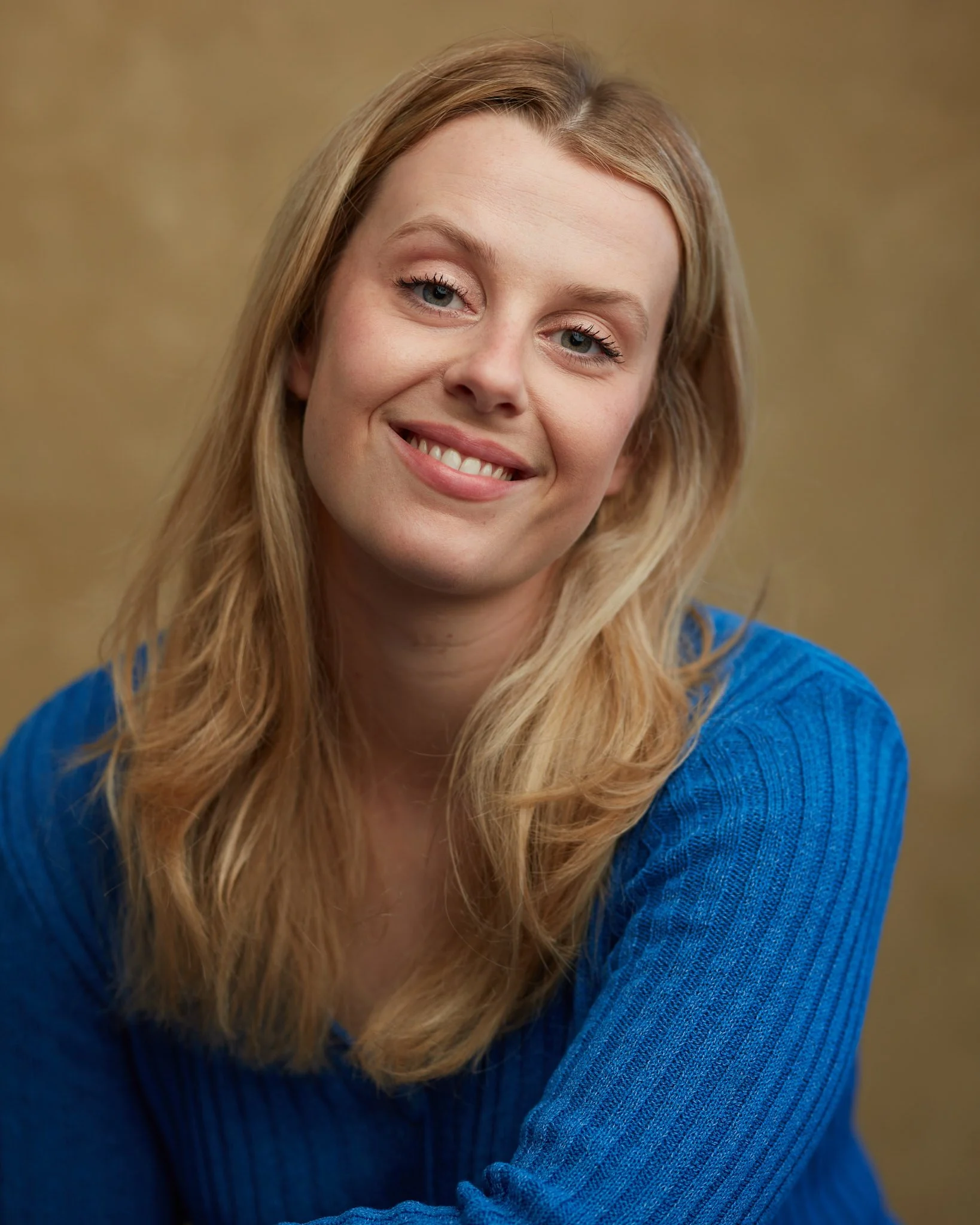 Close-up of a woman smiling, with blonde hair, wearing a blue sweater, against a neutral background.