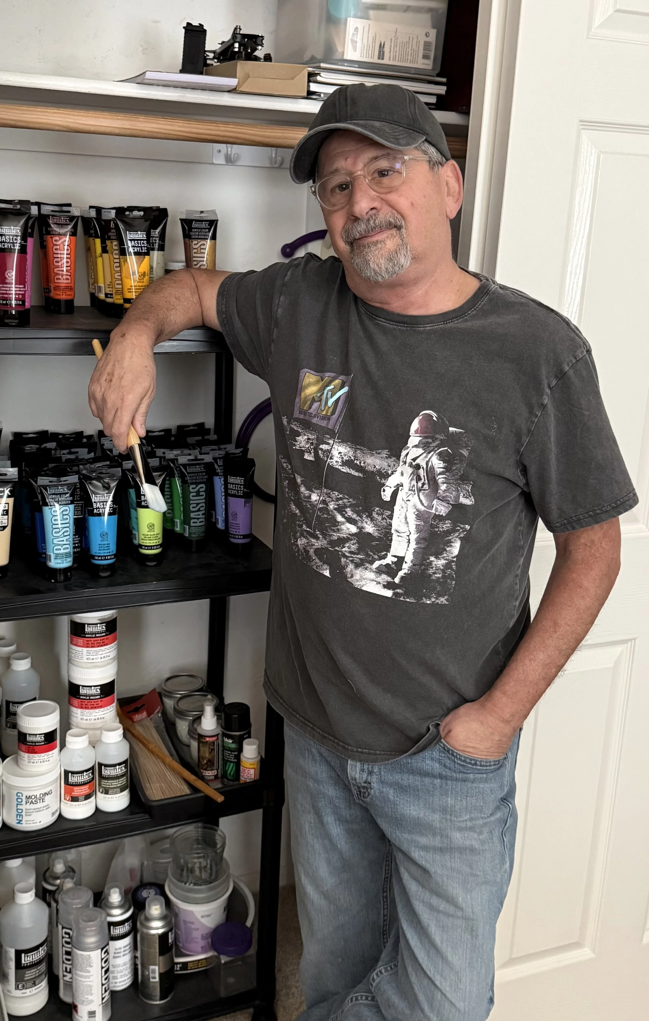 A man wearing glasses, a black cap, a dark gray T-shirt with a space-themed graphic, and jeans stands in a room with shelves of art supplies, including paints, brushes, and bottles.