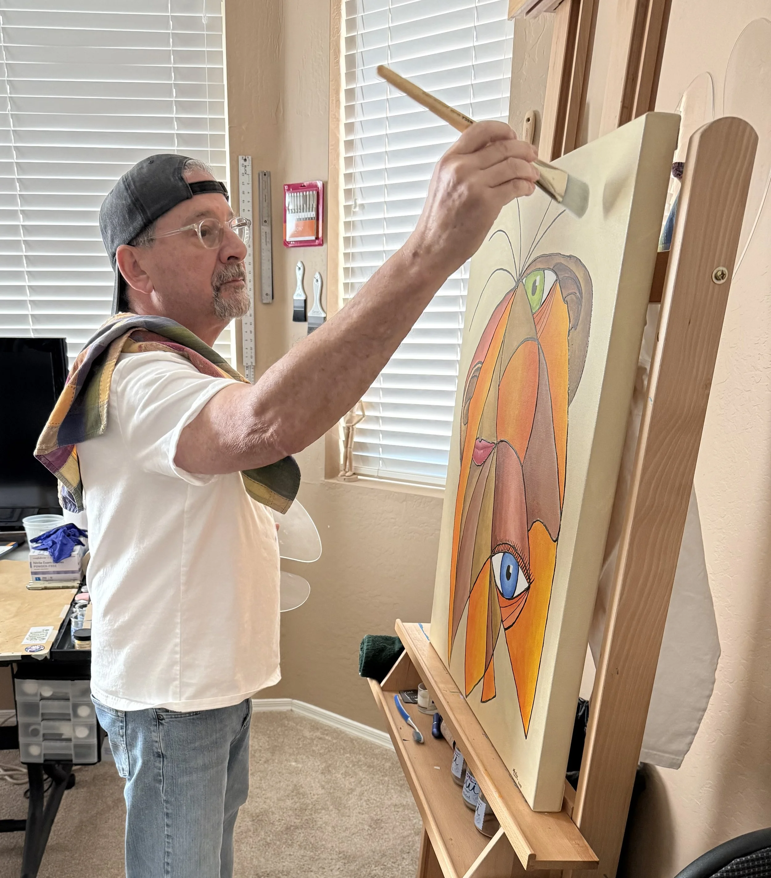 A man with gray hair, glasses, and a black cap is painting a colorful abstract face on a canvas set on an easel. He is using a broad brush and standing in a room with a window covered by horizontal blinds.