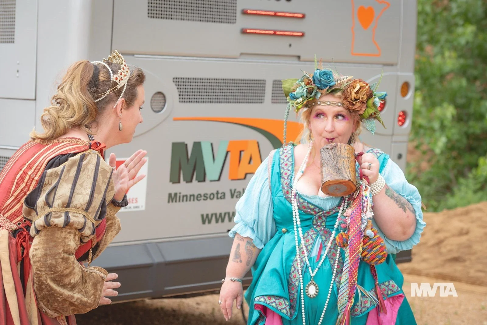Two women dressed in colorful, eccentric costumes are outdoors in front of a Minnesota Valley Transit Authority bus. One woman, with a tiara and vintage-style dress, is smiling and gesturing with her hand. The other woman, wearing a large floral headpiece, colorful dress, and lots of necklaces, is sipping from a mug made from a tree trunk.
