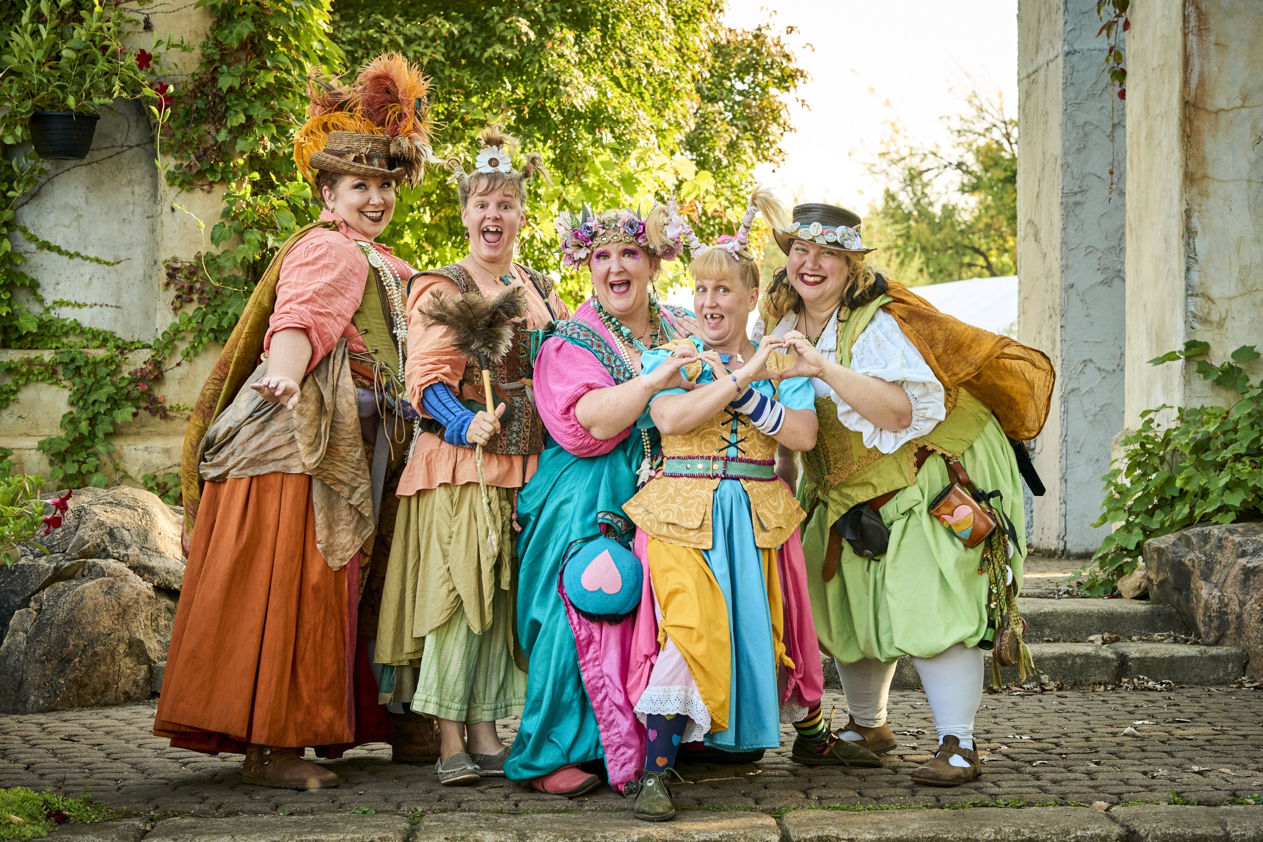 Group of five women dressed in colorful, whimsical outfits with fantasy-inspired accessories, posing outdoors among greenery and stone structures.