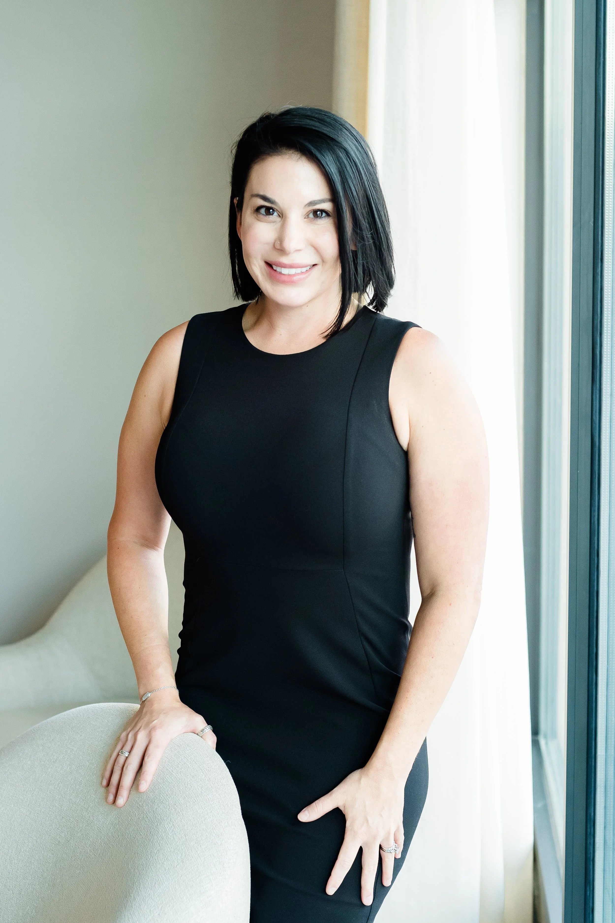 A woman with short black hair and a black sleeveless dress standing indoors near a window, smiling and resting her hand on a beige chair.