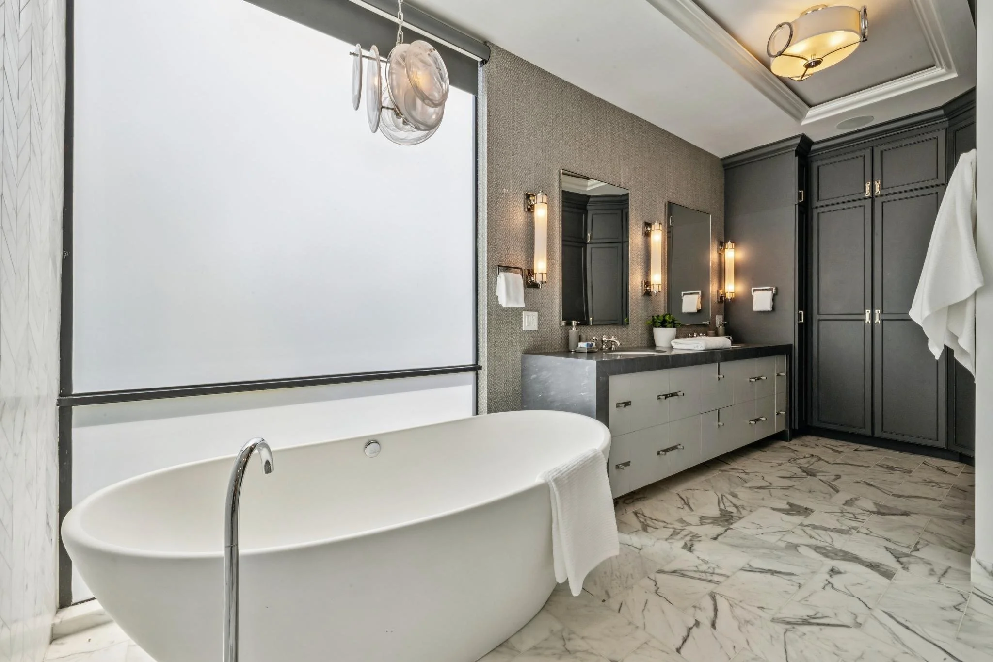 Bathroom with a freestanding bathtub, marble flooring, a vanity with two mirrors, dark cabinetry, and modern lighting fixtures.