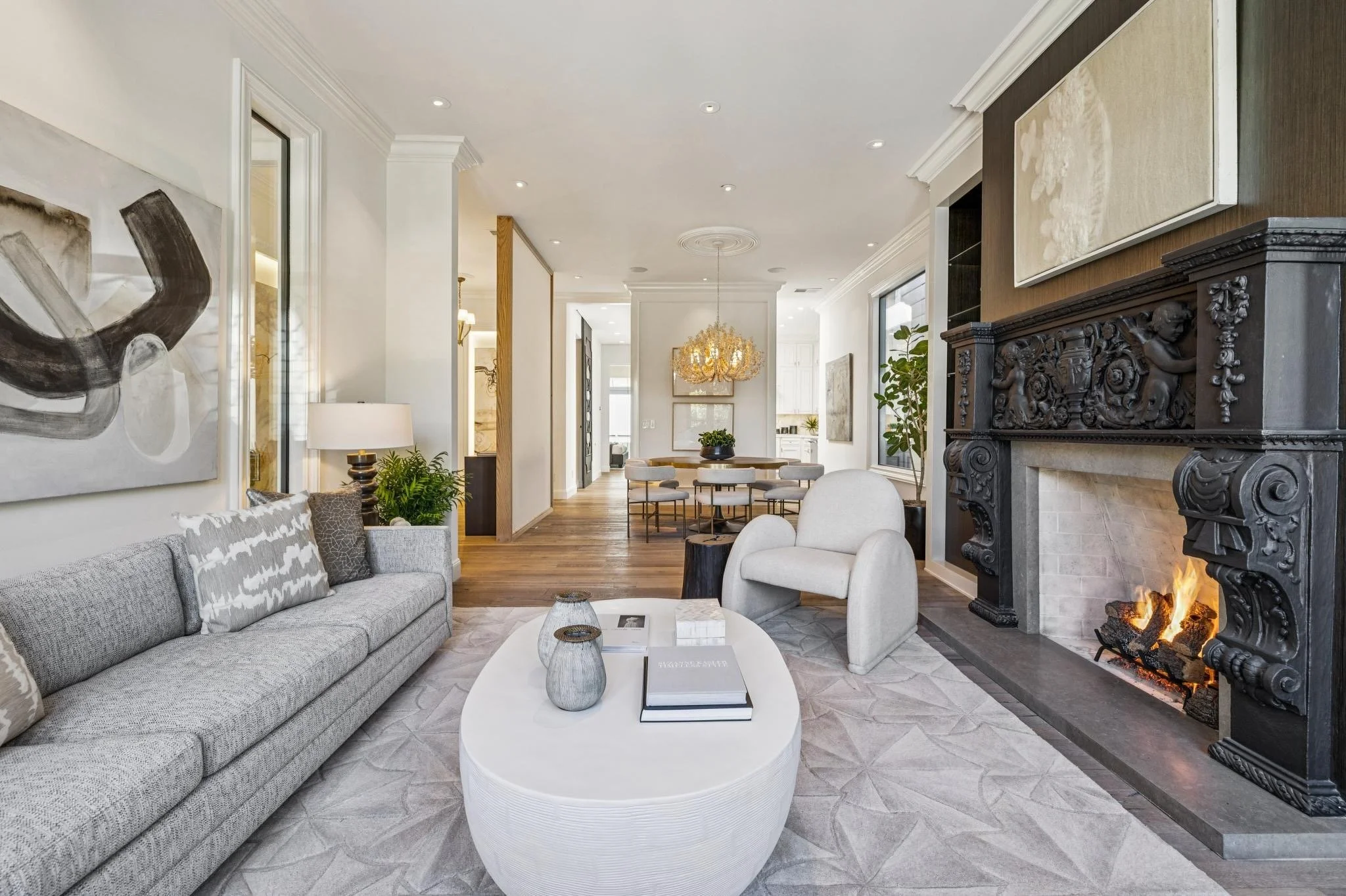Modern living room with gray sofa, white armchair, fireplace with ornate black mantel, artwork on walls, chandelier, and dining area in background.