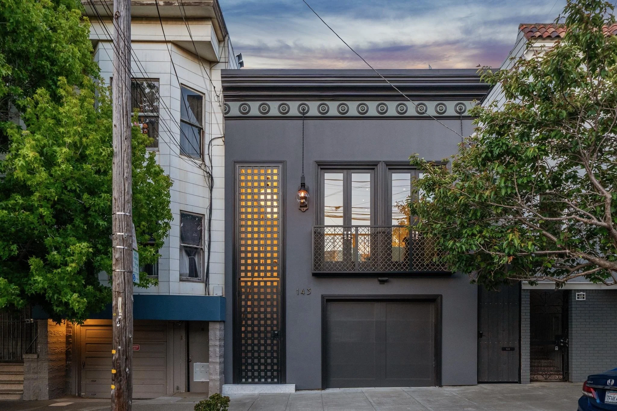 A modern gray house with a unique black-front door featuring a decorative side panel with circular cutouts. The house has two large windows, one on each side of the door. The house number, 143, is visible to the right of the door. Surrounding trees p