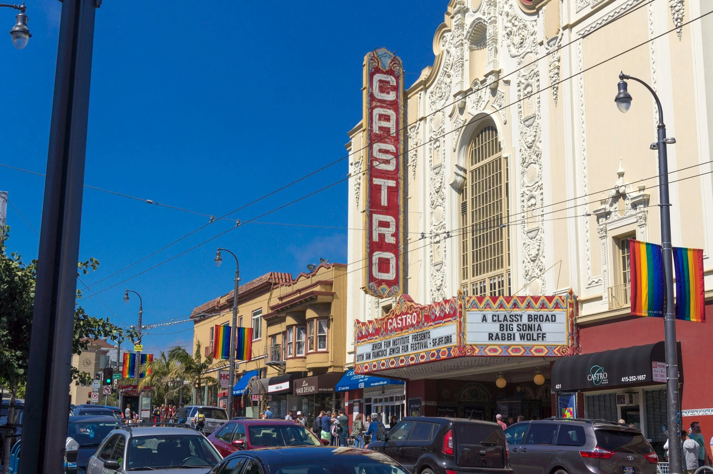 The Castro Theatre on Castro Street with a bright marquee advertising a show called 'A Classy Broad' starring Big Sonia and Rabbi Wolff, with rainbow pride flags hanging outside and a busy street scene with cars and pedestrians.