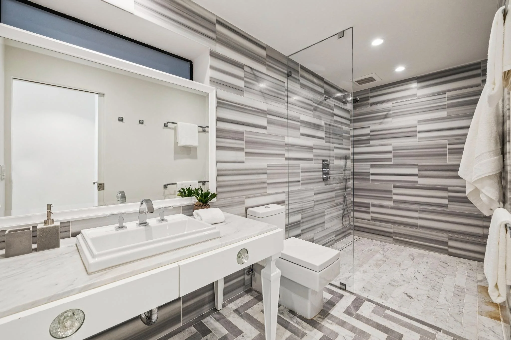 Modern bathroom with a double sink vanity, a toilet, a walk-in shower with a glass door, gray and white striped wall tiles, and a mirror above the sinks.