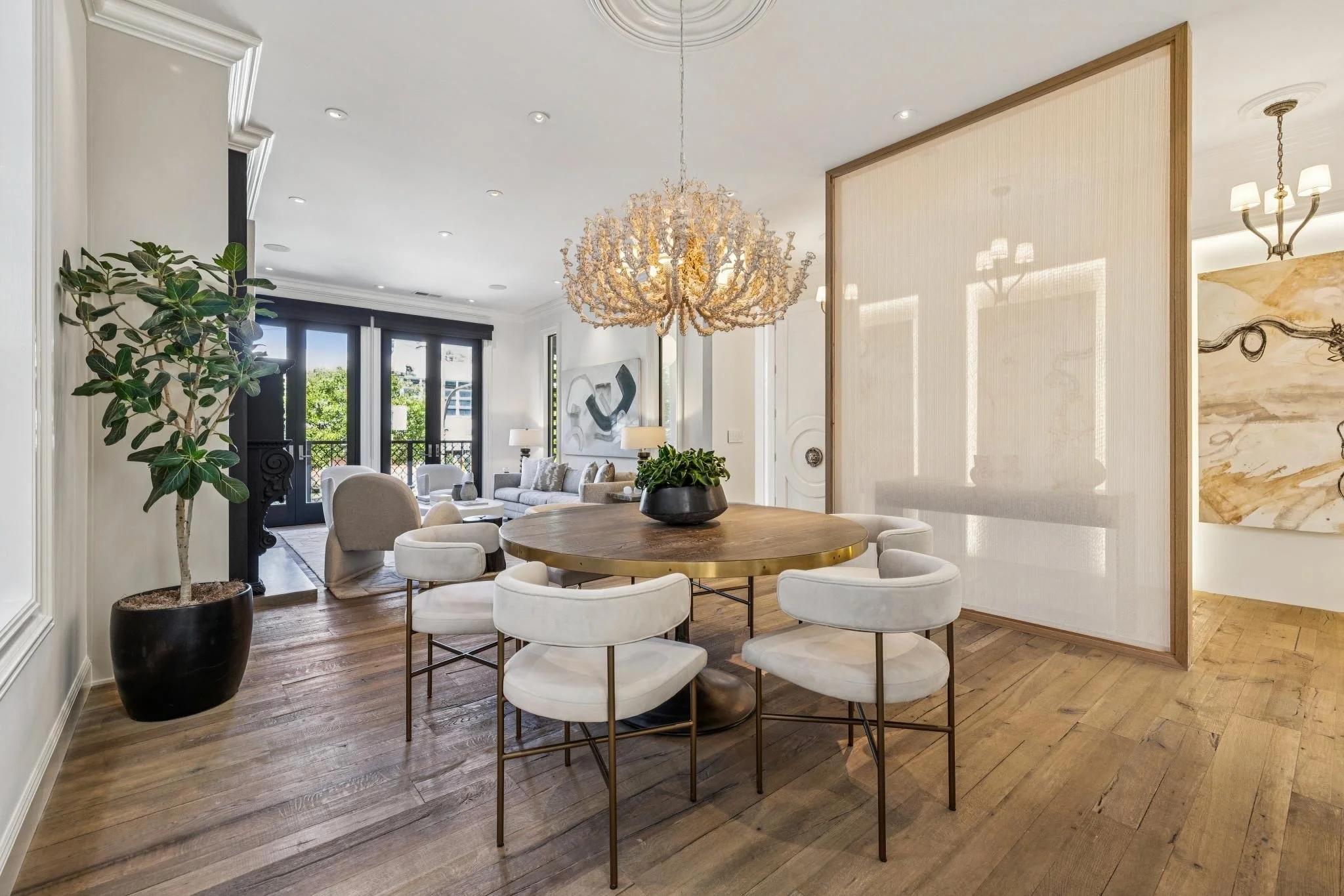 Interior of a modern living and dining area with a round wooden dining table surrounded by white upholstered chairs, a large decorative chandelier, green potted plant, and large windows with a balcony in the background.