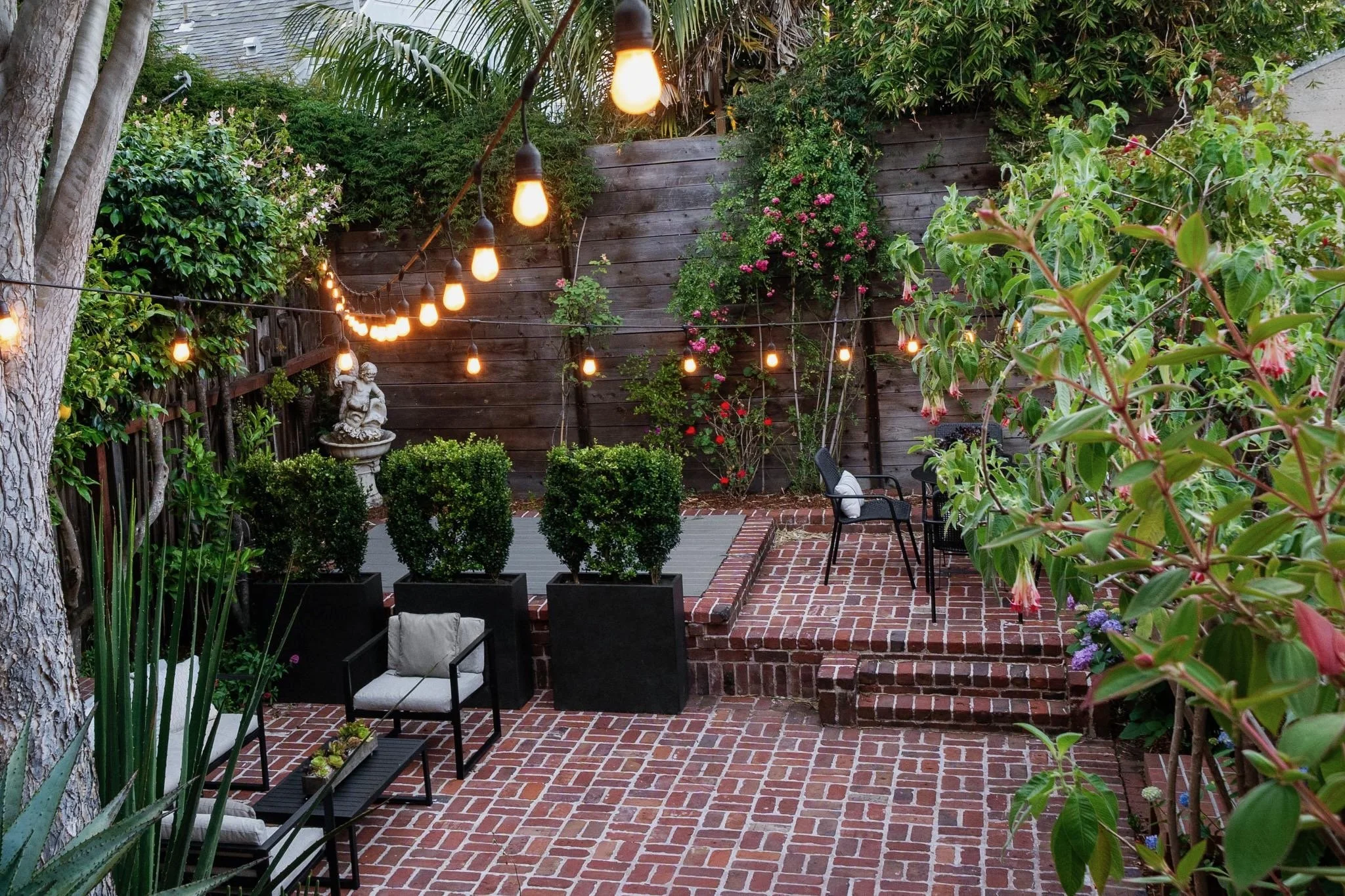 A cozy backyard patio with string lights, lush greenery, potted plants, a stone statue, and outdoor seating on a brick and stone patio.