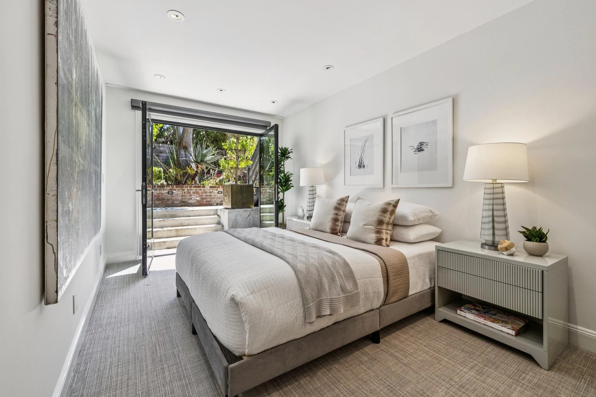 A modern bedroom with a large bed, neutral-colored bedding, and decorative pillows. There's a white bedside table with a striped lamp, a small plant, and some decorative stones. Two framed black-and-white wall art pieces hang above the bed. An open glass door leads to a small outdoor patio with brick stairs, greenery, and plants.