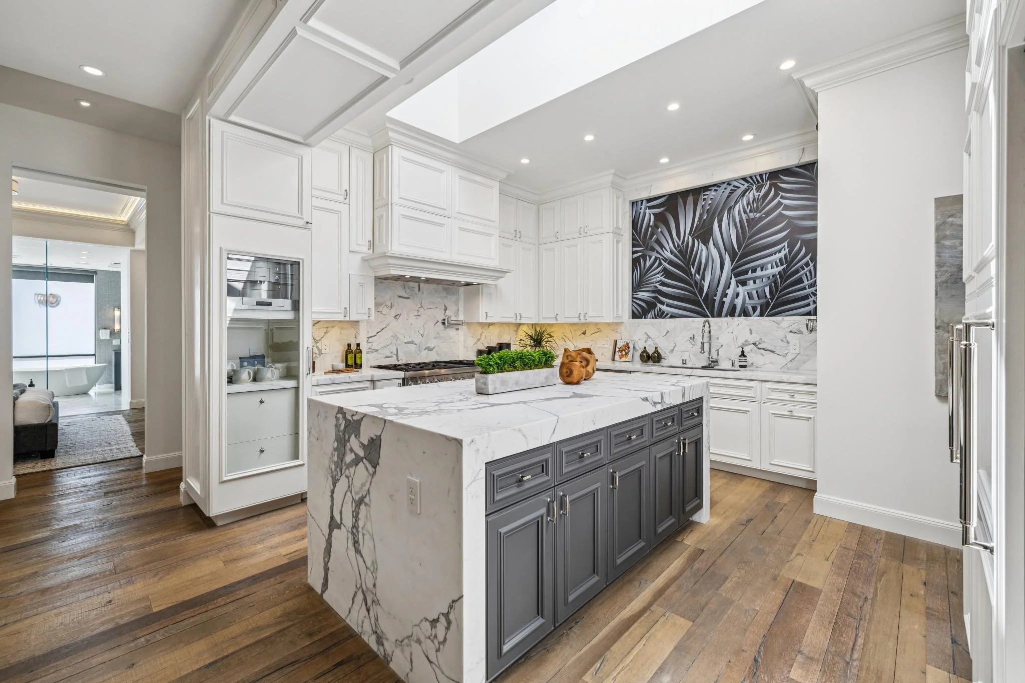 A modern kitchen with white cabinets, a marble island with a dark gray base, and a black and white tropical leaf mural on the wall.
