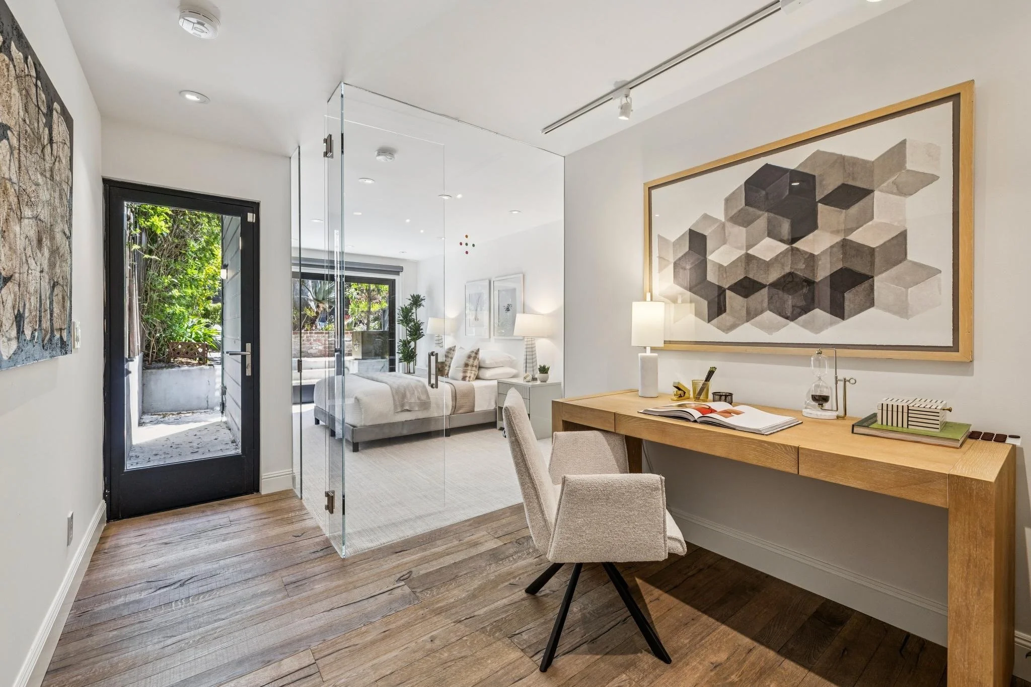 Interior view of a modern house with a small workspace and a bedroom visible through a glass partition. Wood flooring, white walls, large wall art, and plants near the back door.