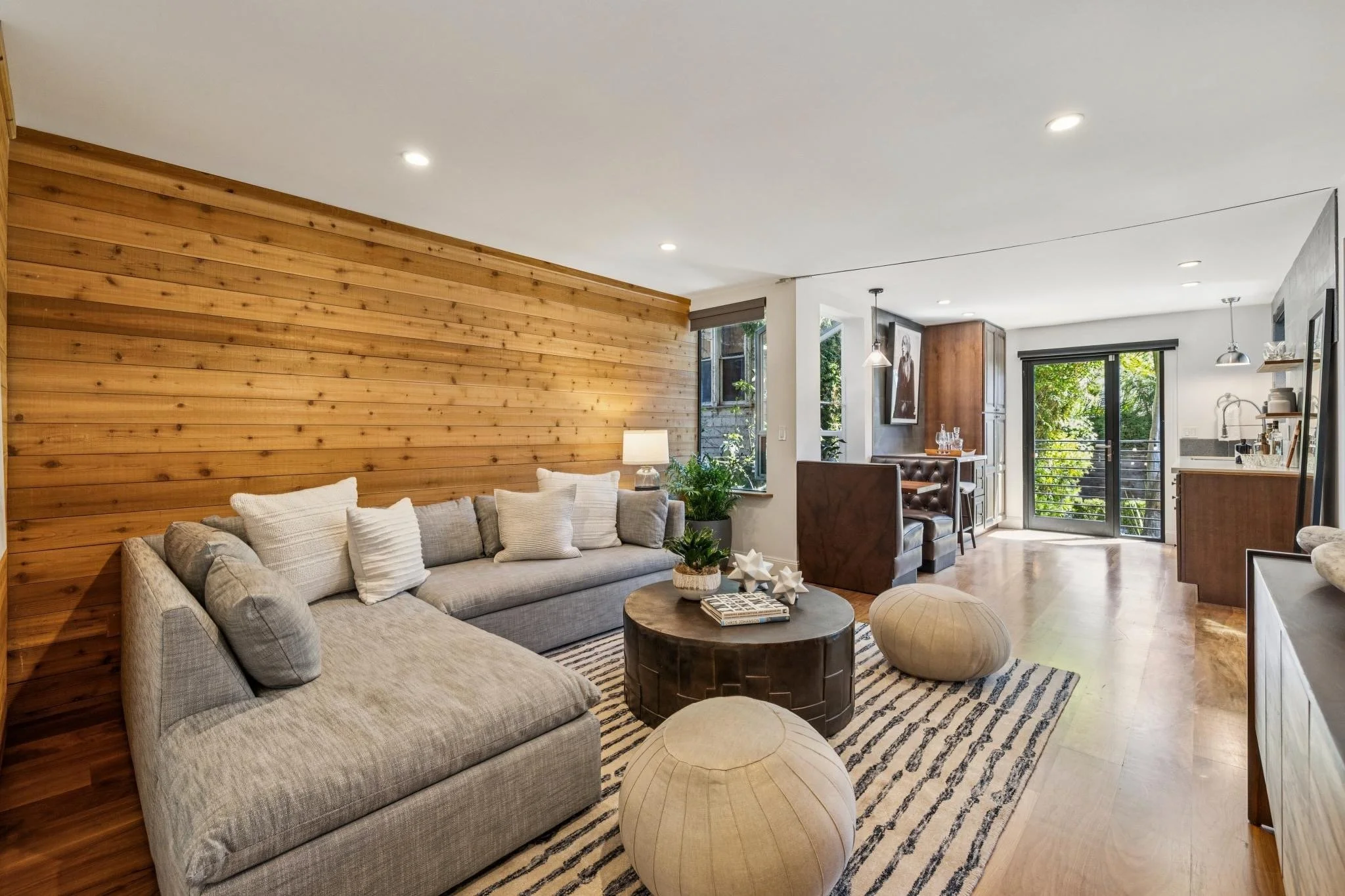 Living room with a light gray sectional sofa, wooden accent wall, round coffee table, and decorative pillows. Kitchen and dining area with dark cabinetry, sliding glass door leading outdoors, and modern lighting.
