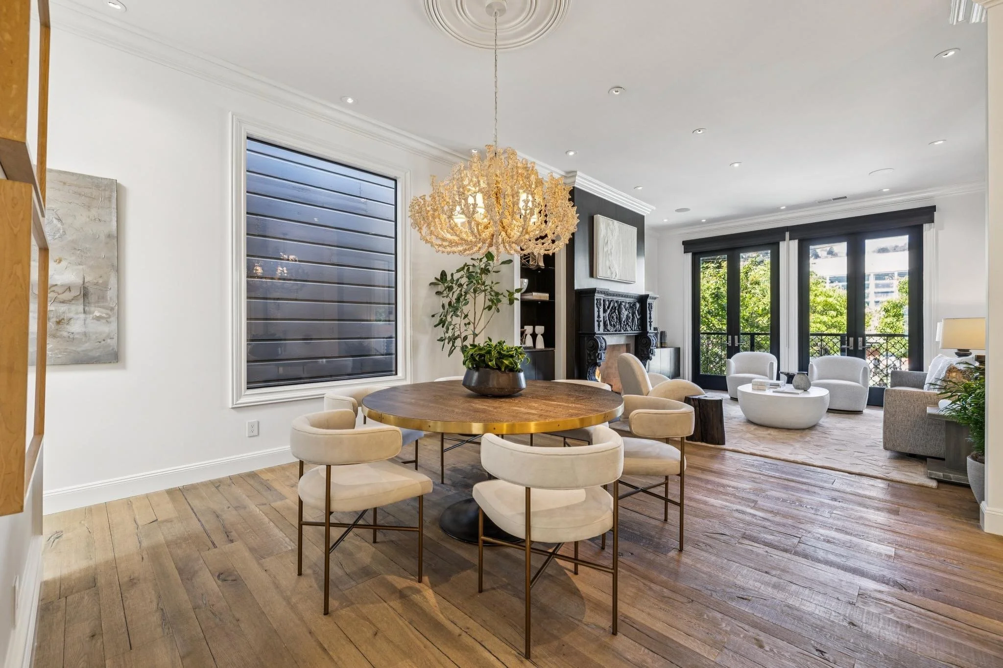 A spacious living and dining room with a round wooden table surrounded by beige upholstered chairs, a white sofa set, large windows with black frames, a chandelier hanging above the table, hardwood flooring, and a decorative dark fireplace with white abstract artwork above it.