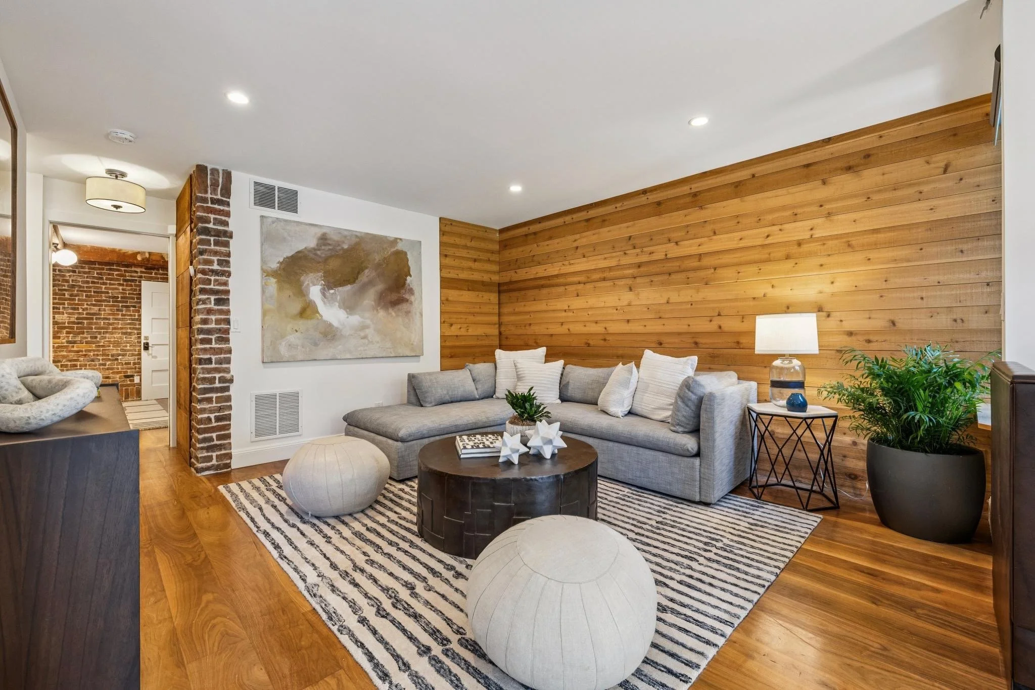 Living room with a gray sectional sofa, a black and white striped rug, a round wooden coffee table with decorative objects, beige poufs, a large abstract painting, a wooden wall panel, potted plants, and ceiling lights.