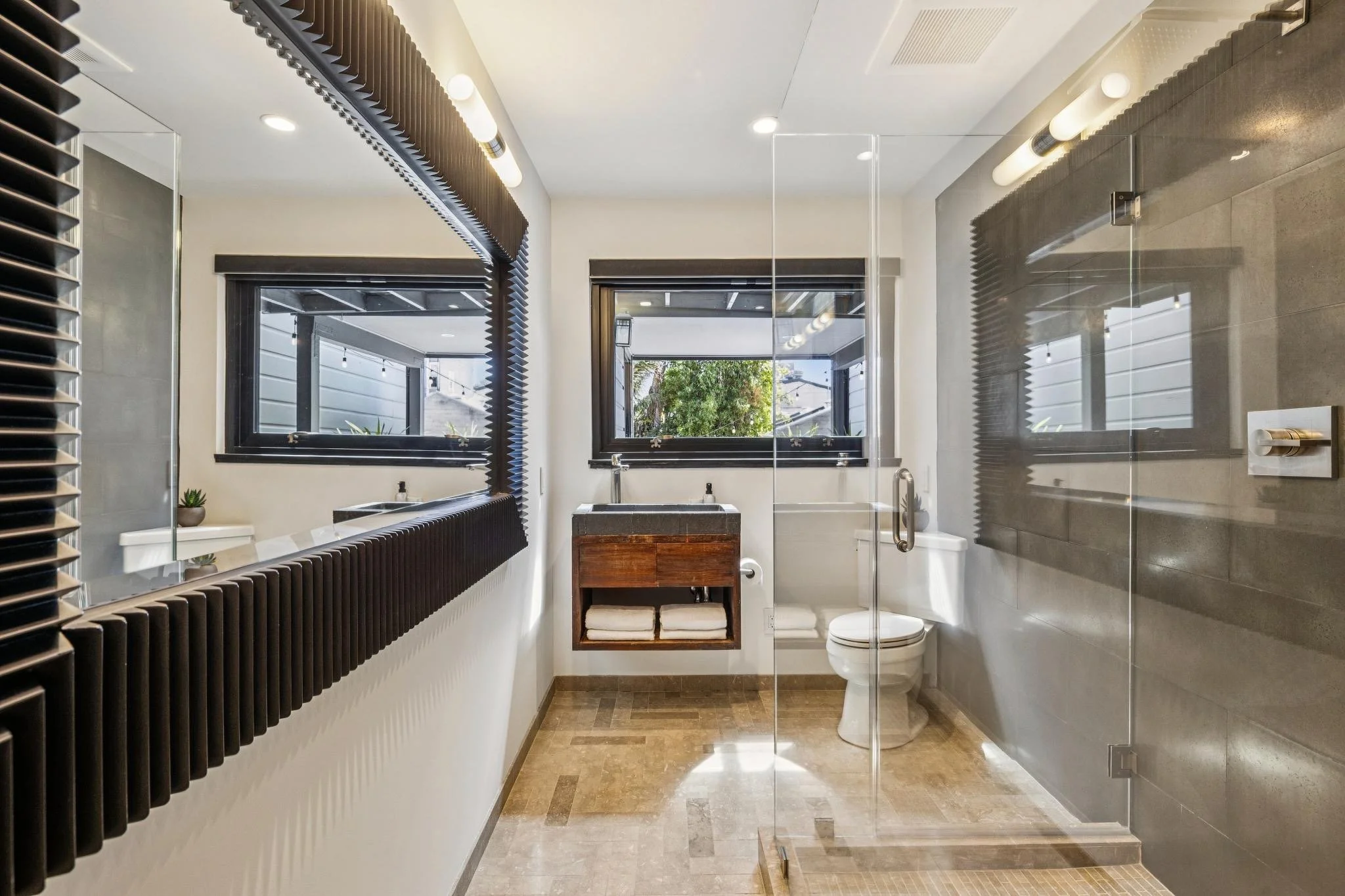Modern bathroom with a large window, black accents, a walk-in shower with glass wall, white toilet, small wooden storage, and ceramic floor tiles.