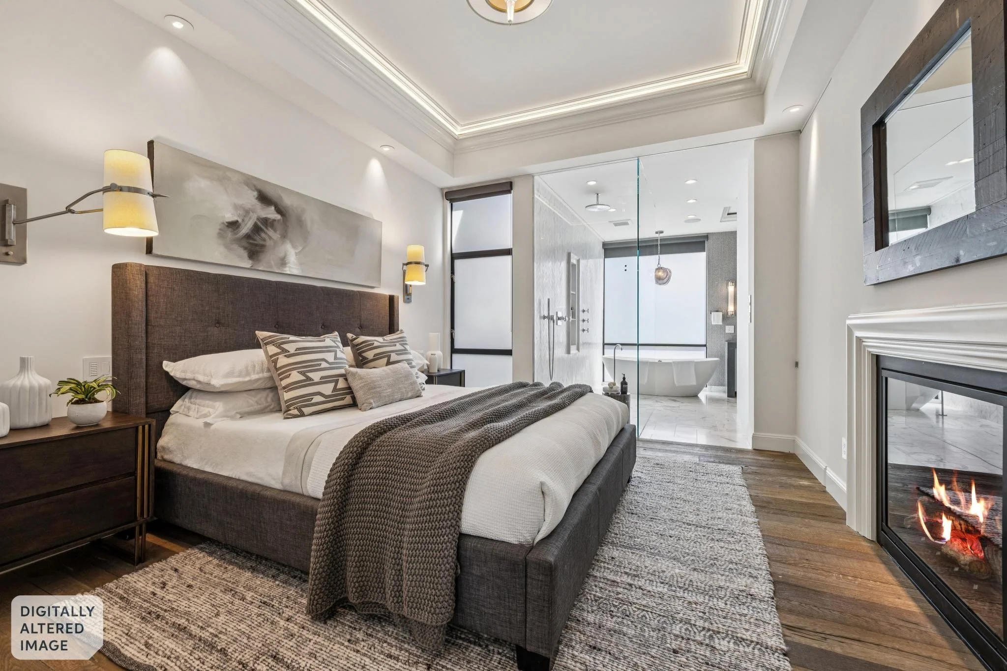 Modern bedroom with a grey upholstered bed, patterned pillows, and a knitted blanket. There is a nightstand with decorative items, wall-mounted lamps, and abstract artwork. The room opens to a bathroom with a freestanding bathtub, glass shower enclos
