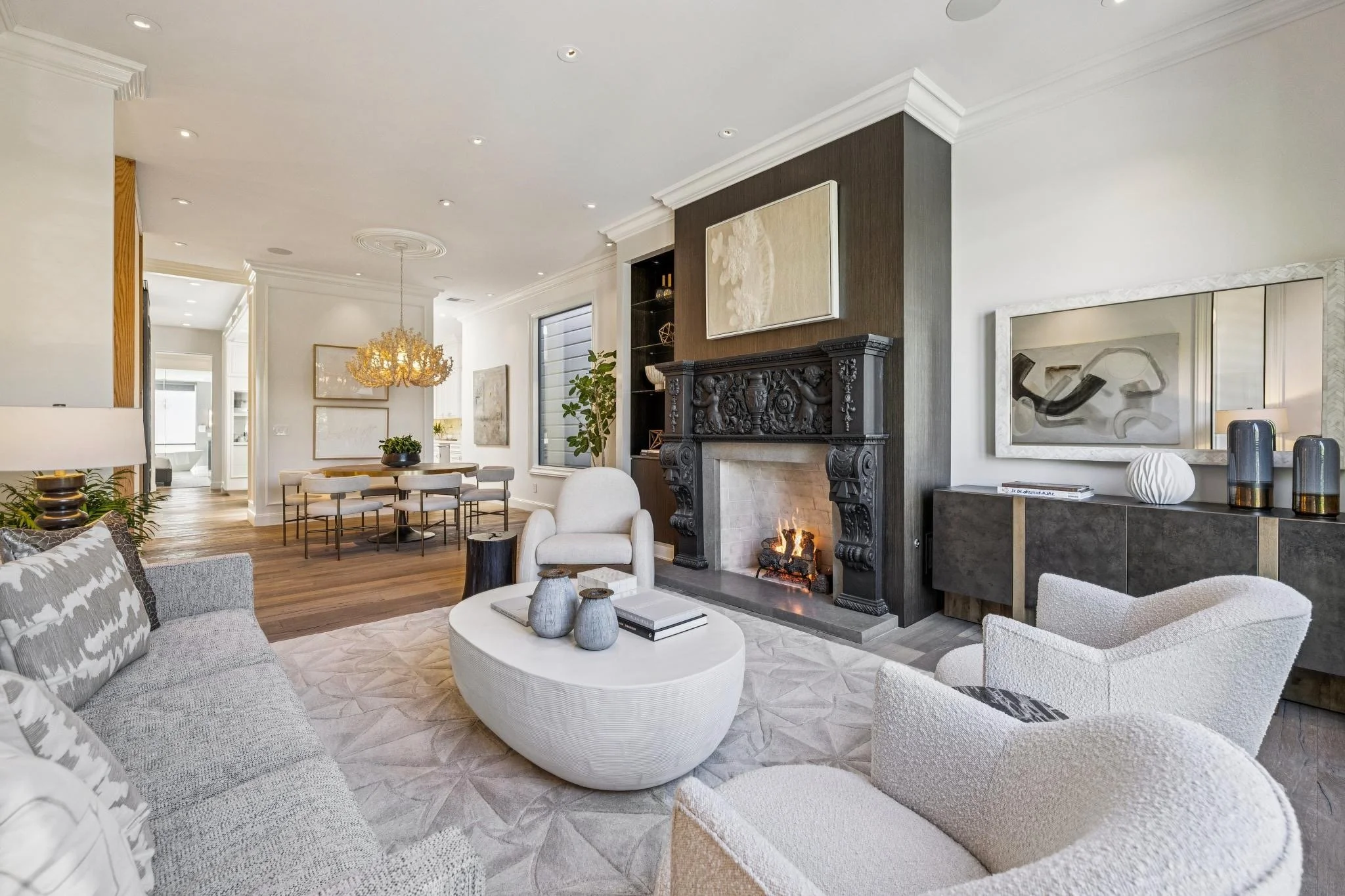 A spacious, modern living room with a white and gray color scheme, featuring a fireplace with black ornate detailing, contemporary artwork, a white armchair, a round coffee table with decorative vases, a beige patterned sofa, and a dining area with a wooden table and white chairs. The room has hardwood flooring, crown molding, and recessed lighting, with accents like a gold chandelier and potted plants.