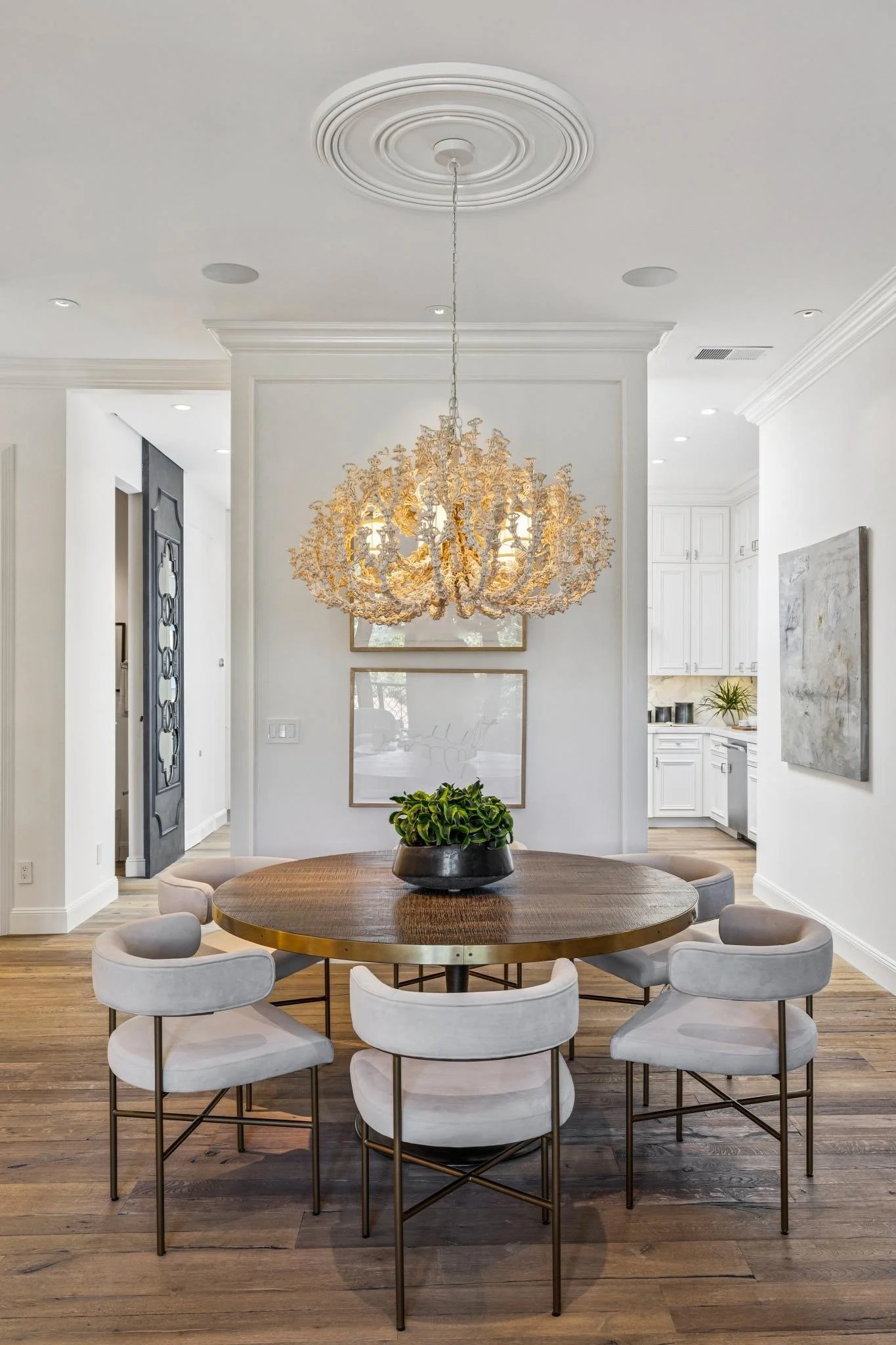 Dining room with a round wooden table surrounded by eight white upholstered chairs, a black bowl with a green plant, a decorative chandelier, and white walls.