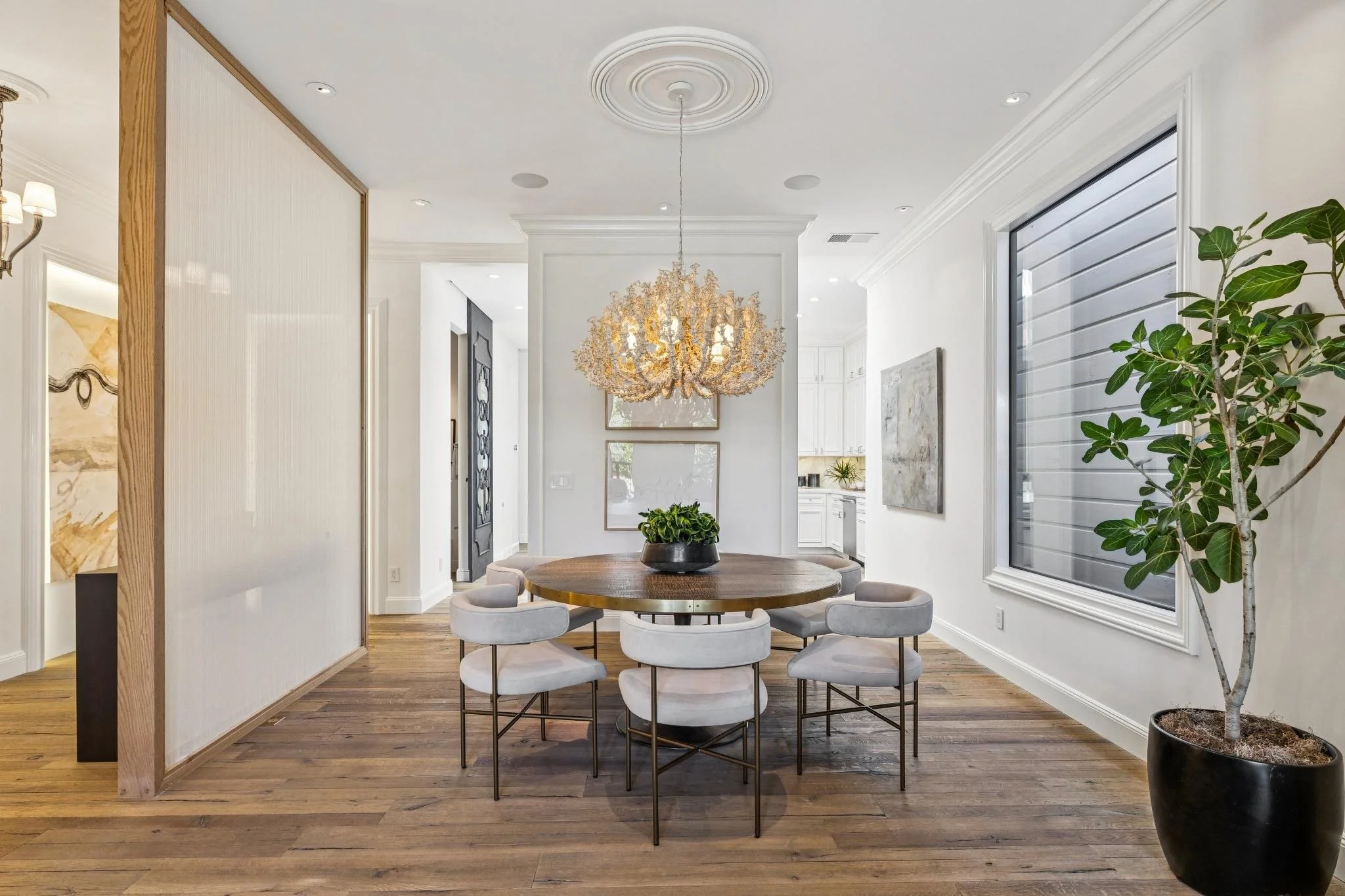 A dining area with a round wooden table surrounded by six upholstered gray chairs, a large chandelier hanging above, a potted plant on the right, and a window with blinds on the right wall. The room has white walls, crown molding, and hardwood floors