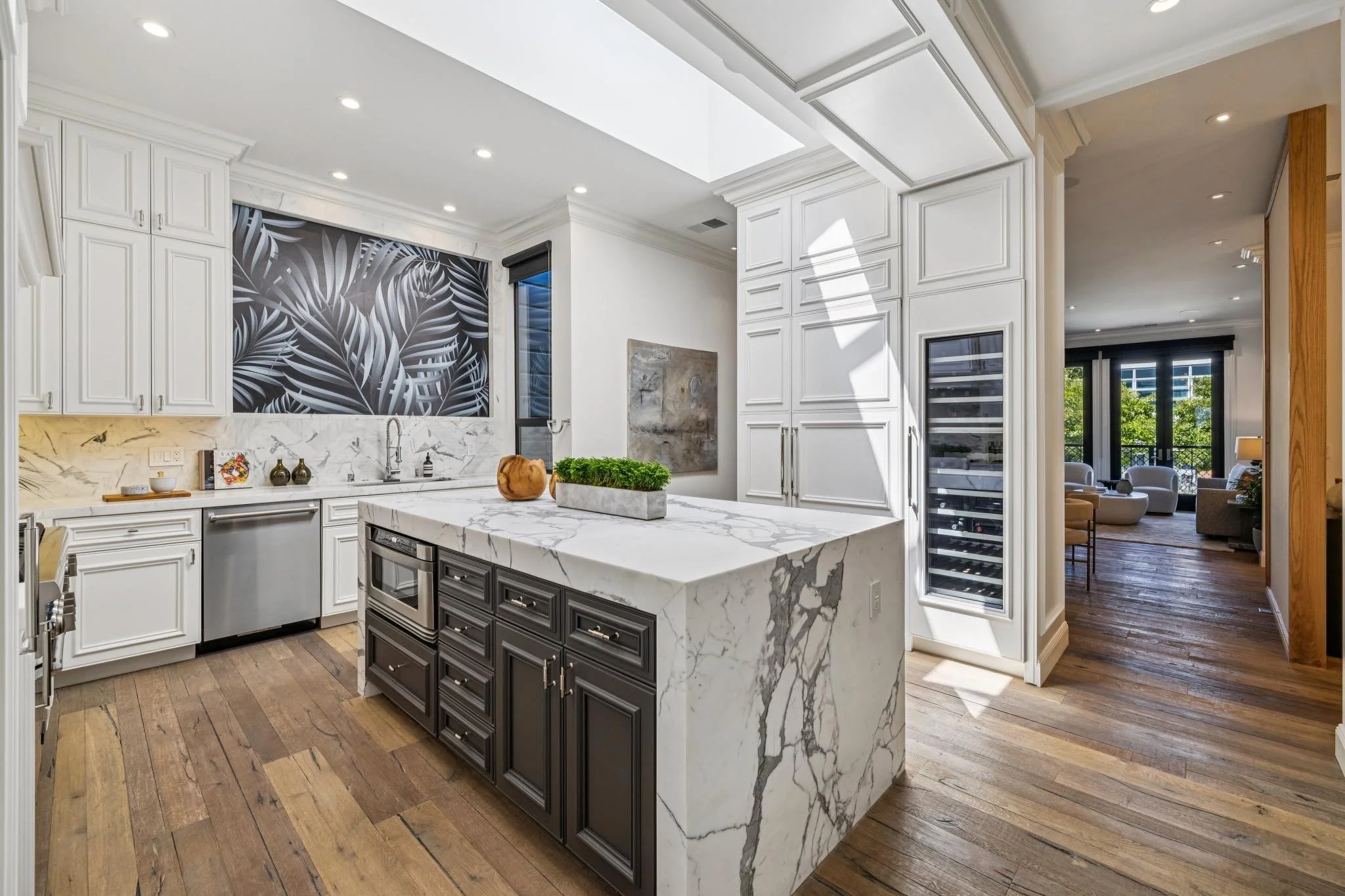 Modern kitchen with white cabinets, black and white marble island, wooden flooring, and a backsplash with a black and white leafy pattern. Opens to a living room with large windows and outdoor view.