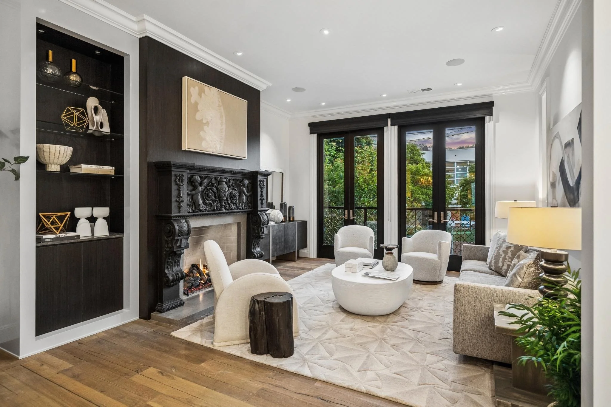 A modern living room with a black fireplace, white armchairs, a round coffee table, large windows showing trees outside, and decorative shelves with art pieces.