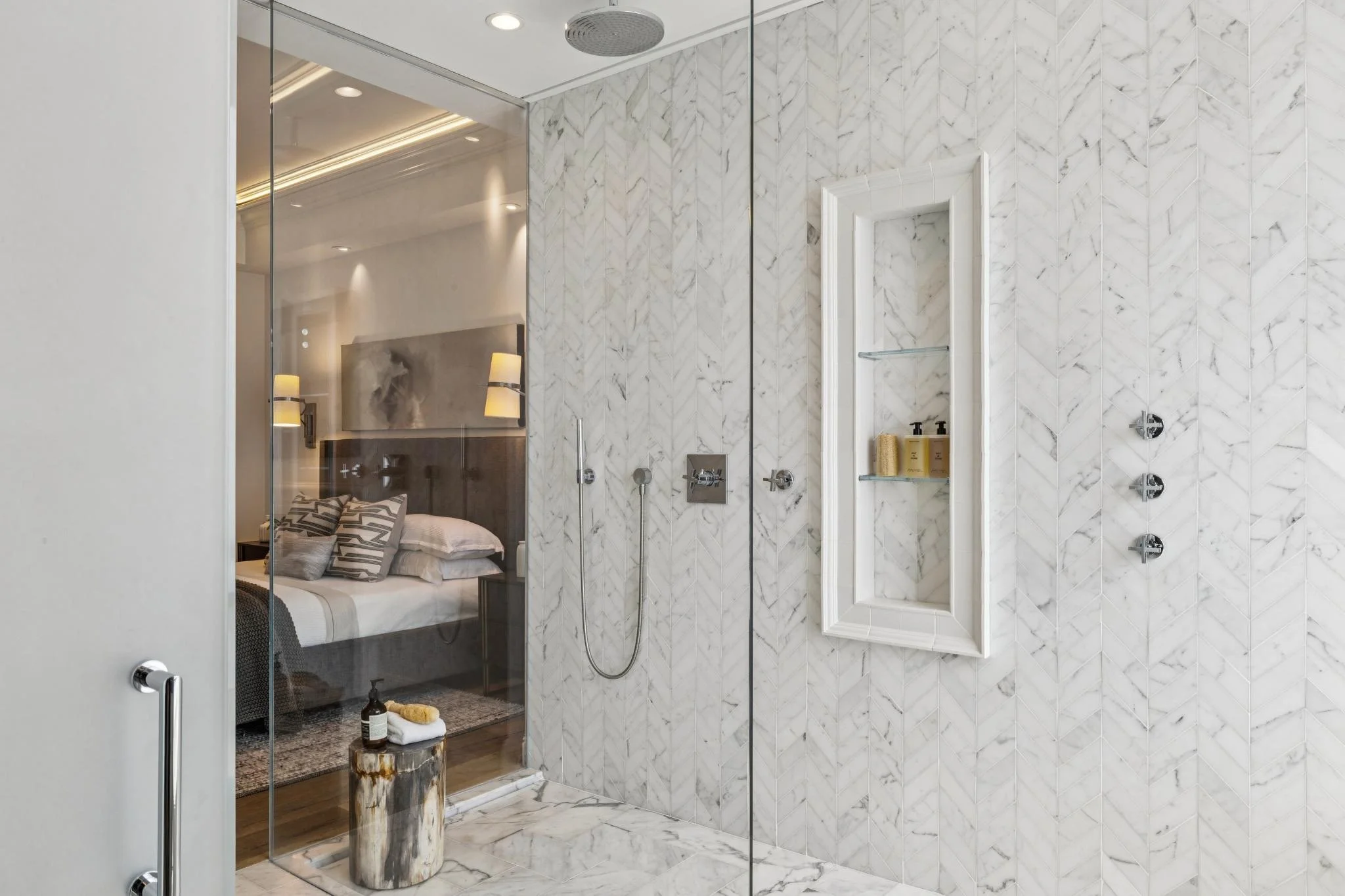 Modern bathroom with white marble walls and flooring, glass shower enclosure, and a walk-in shower with multiple showerheads. Through the glass, a bedroom with bed, pillows, and bedside lamps is visible.