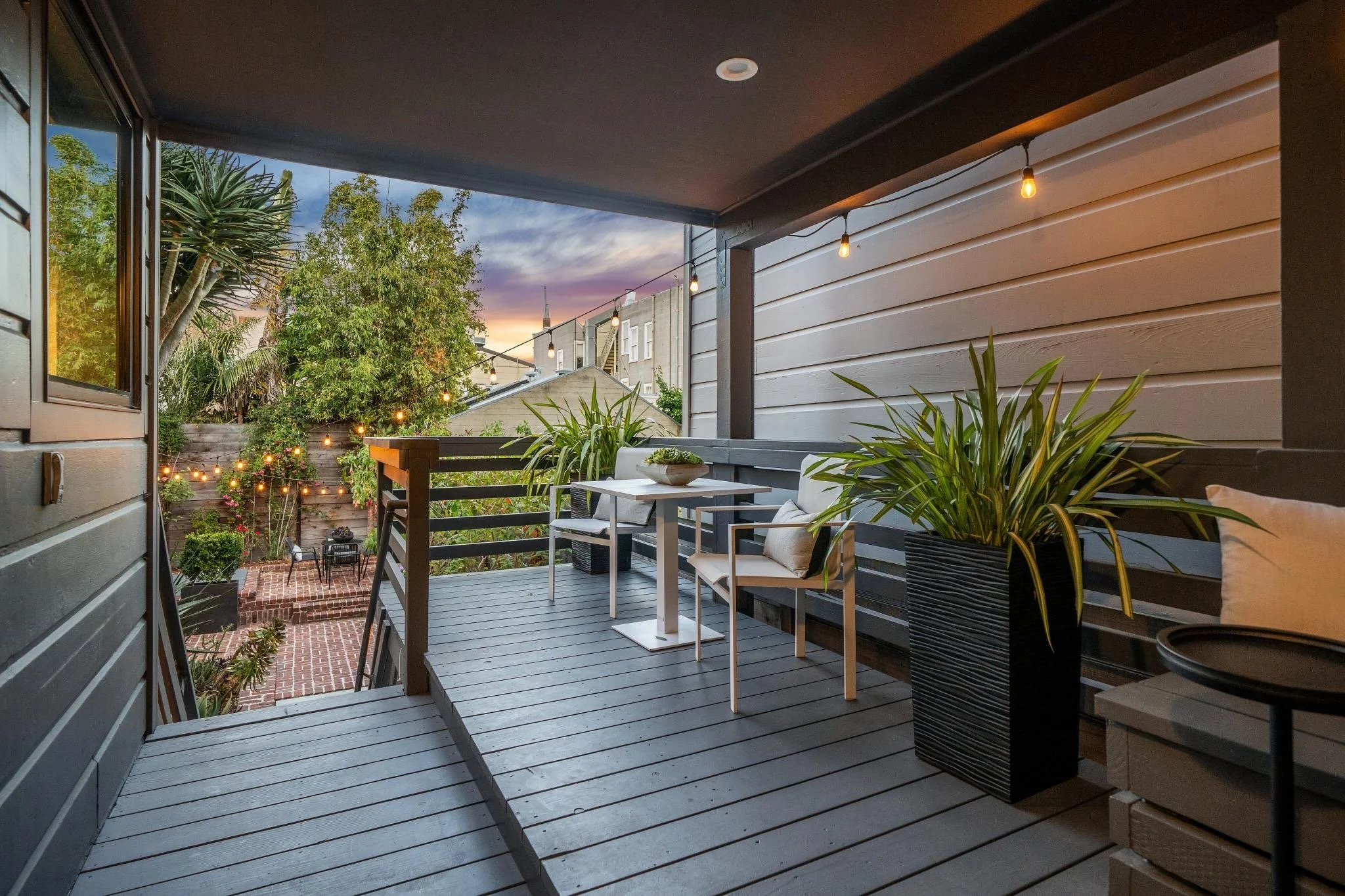 A cozy balcony with gray wooden flooring, outdoor furniture, potted plants, and string lights, overlooking a backyard with trees and a brick patio at sunset.