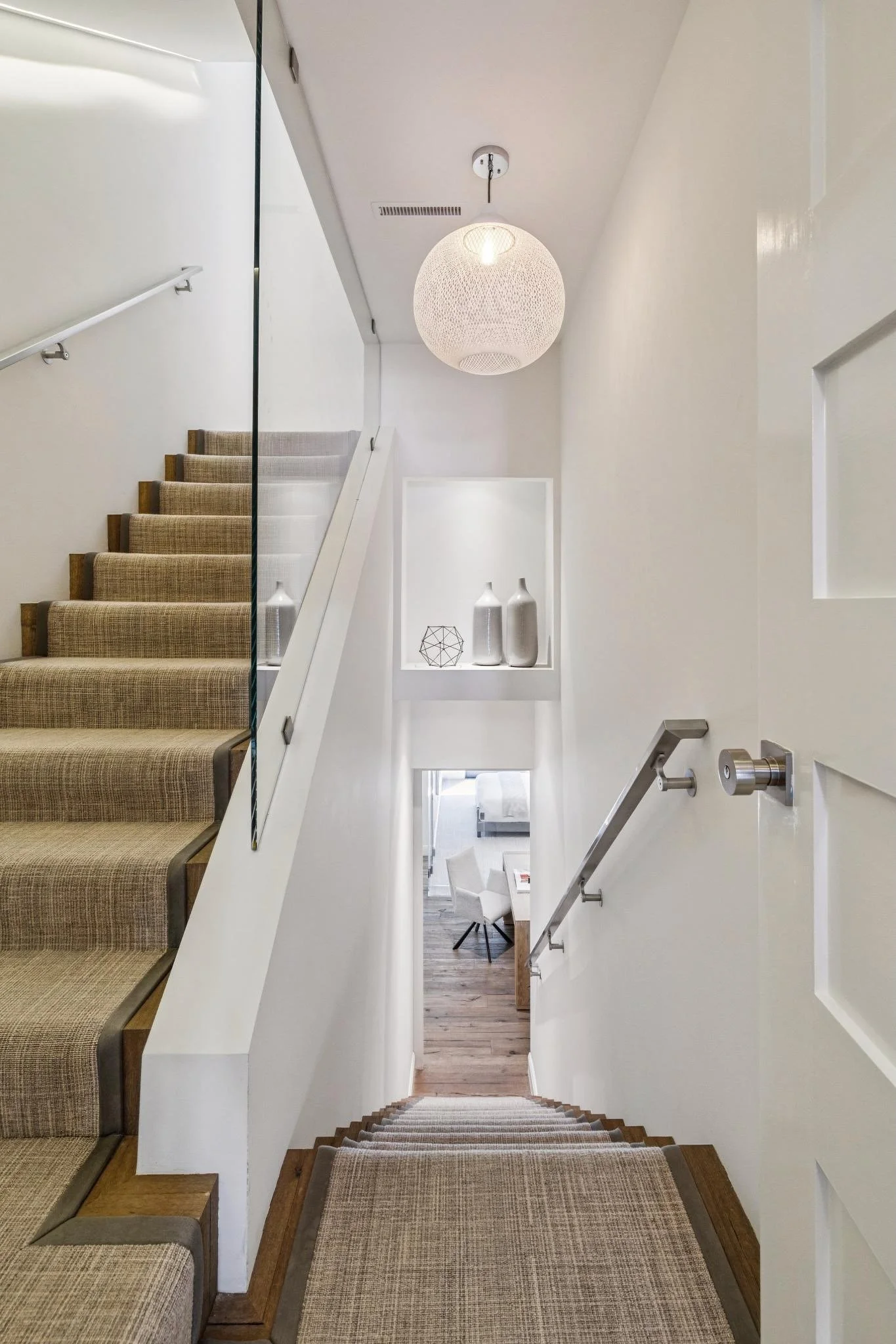 View of indoor staircase with beige carpeted steps, white walls, and modern decor including a white spherical pendant light and decorative vases.