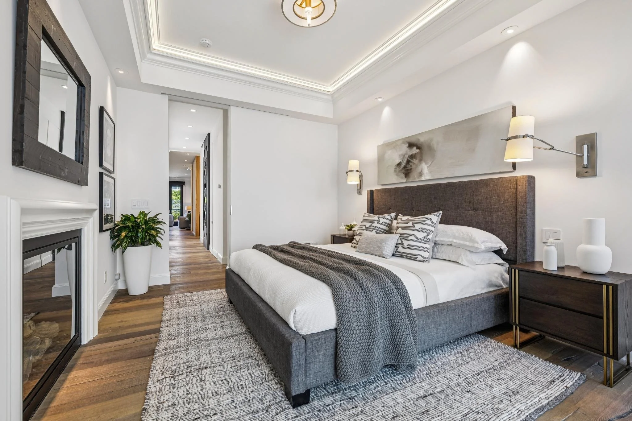 A modern bedroom with a large bed featuring gray headboard, striped pillows, and a gray blanket. Beside the bed are dark wooden nightstands and white vases. The room has white walls, a ceiling with recessed lighting and crown molding, hardwood floors