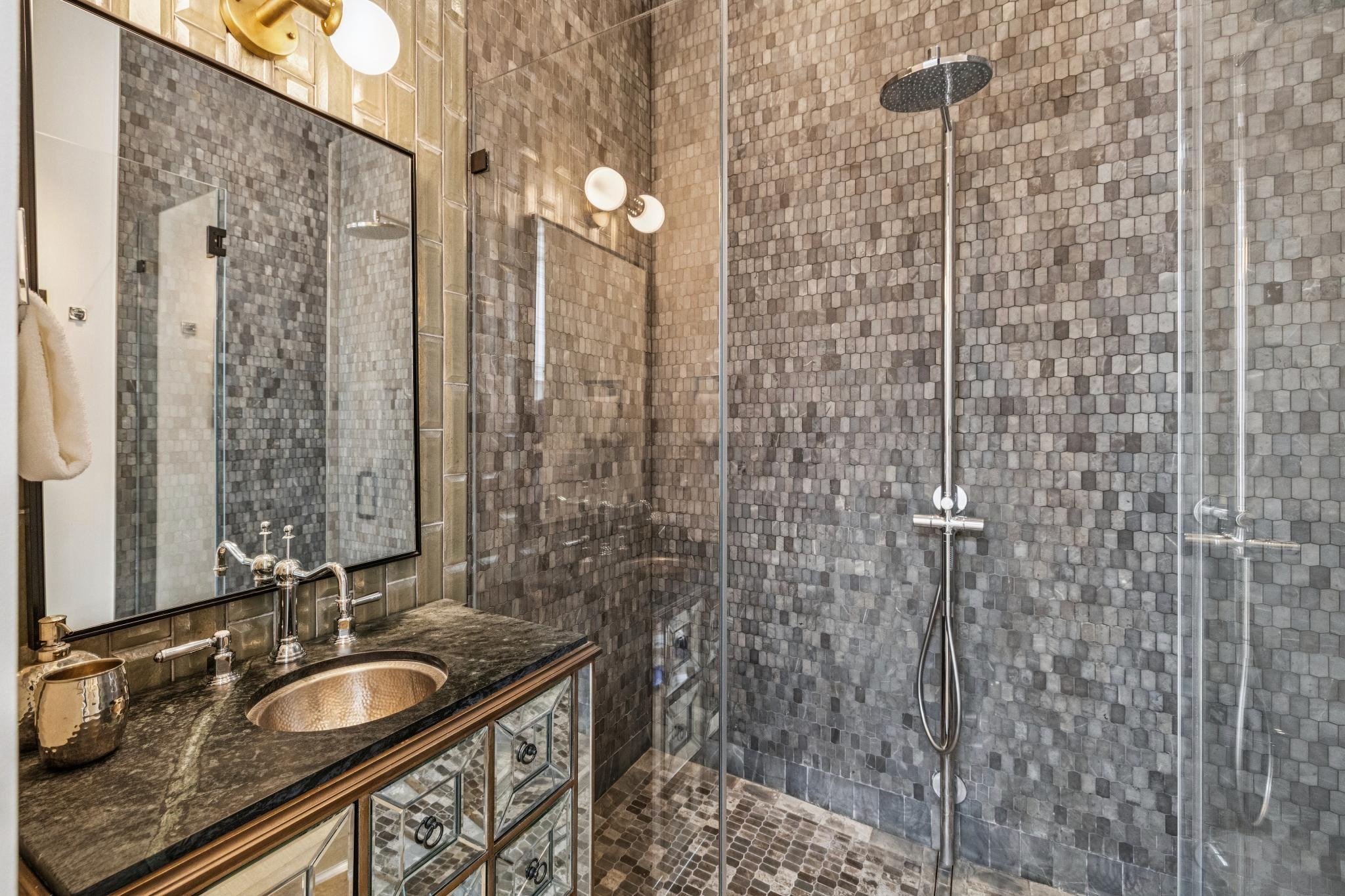 A modern bathroom with gray mosaic tile walls and floor, a glass shower enclosure with a rain showerhead, a black countertop with a gold sink, and a large mirror with vanity lighting.