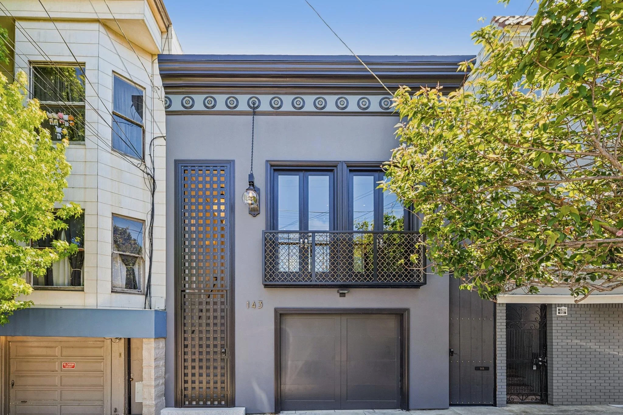 Front of a modern townhouse with gray exterior, a balcony with metal railing, large windows, and a garage door. Houses are adjacent and some greenery is visible.