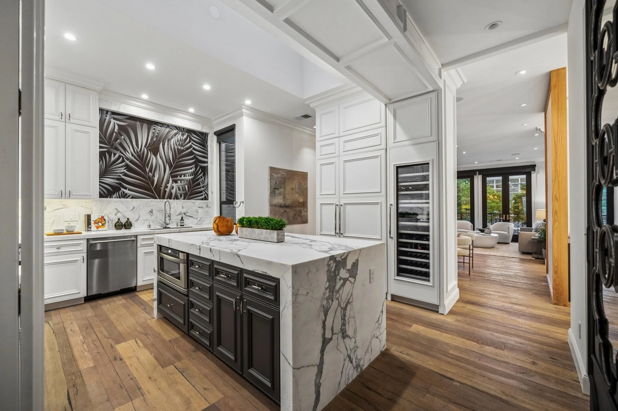 Modern kitchen with white and black cabinetry, marble island, wood flooring, and a view into a living area with large windows and greenery outside.