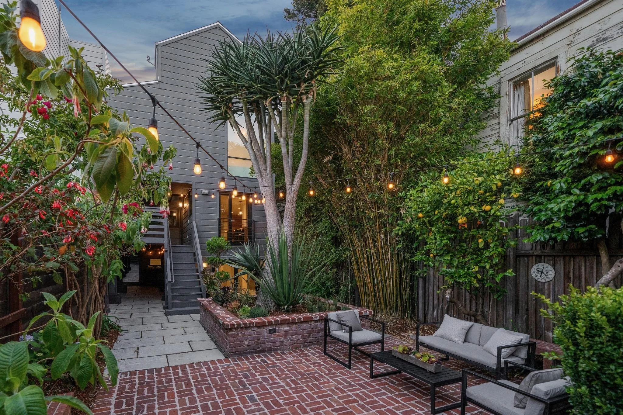 Residential backyard with patio, outdoor seating, string lights, and lush greenery, including trees and flowering plants, during evening hours.