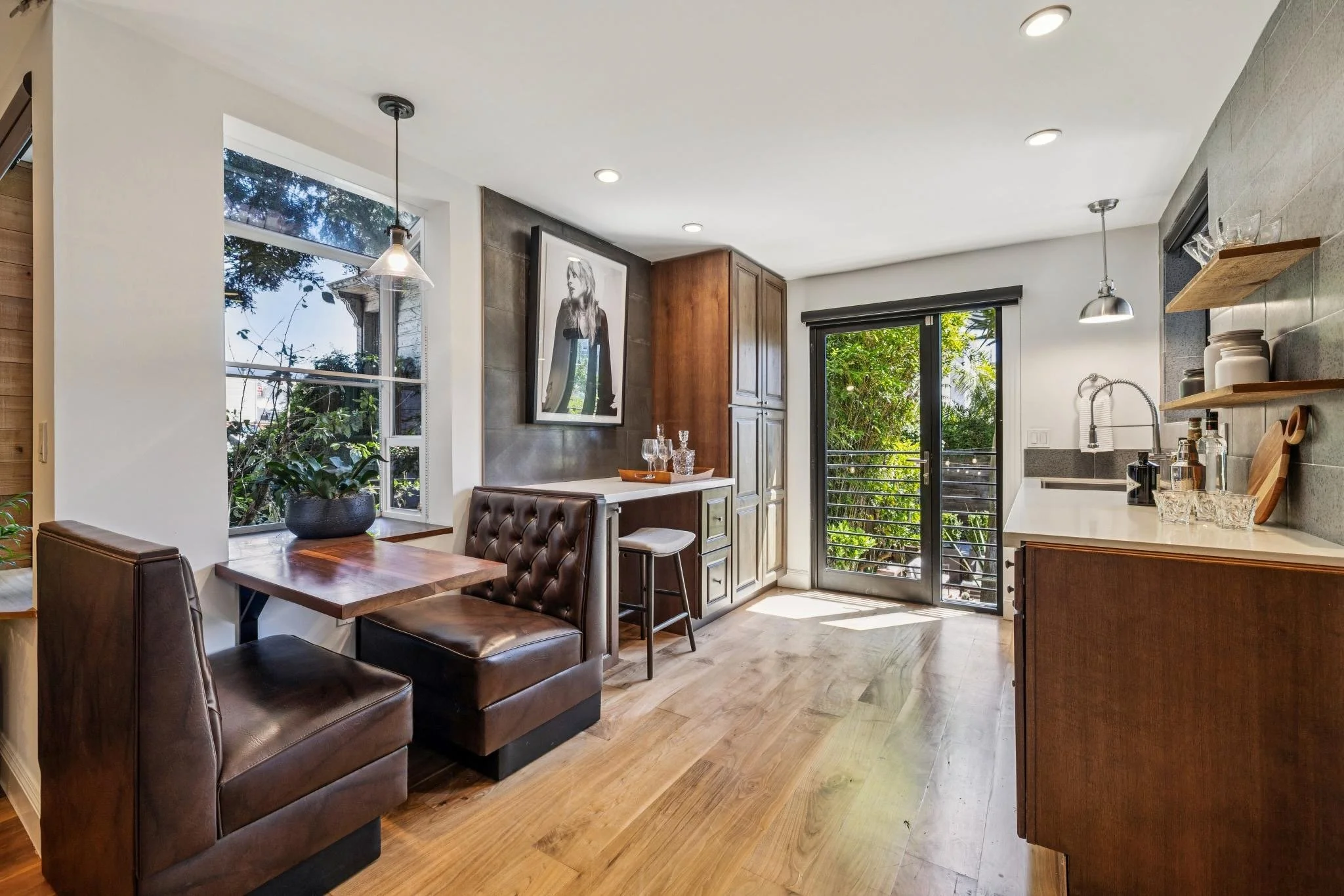 A modern kitchen and dining area with wooden flooring, a small dining table with two leather chairs, a large window showing greenery outside, floating shelves with kitchen supplies, and a black and white portrait on the wall.
