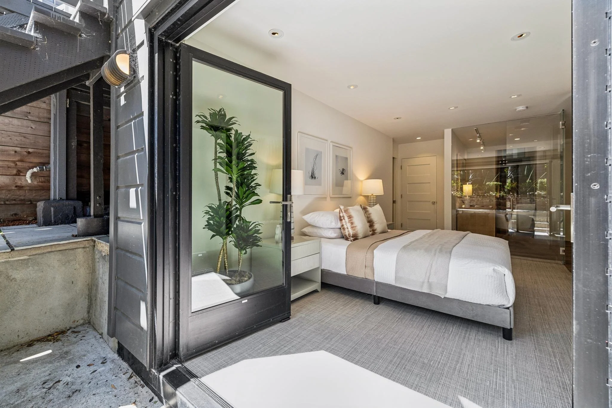 Interior of a modern bedroom with a bed, side table, lamps, artwork on the wall, and a large glass window, seen from an outdoor patio with a metal structure.