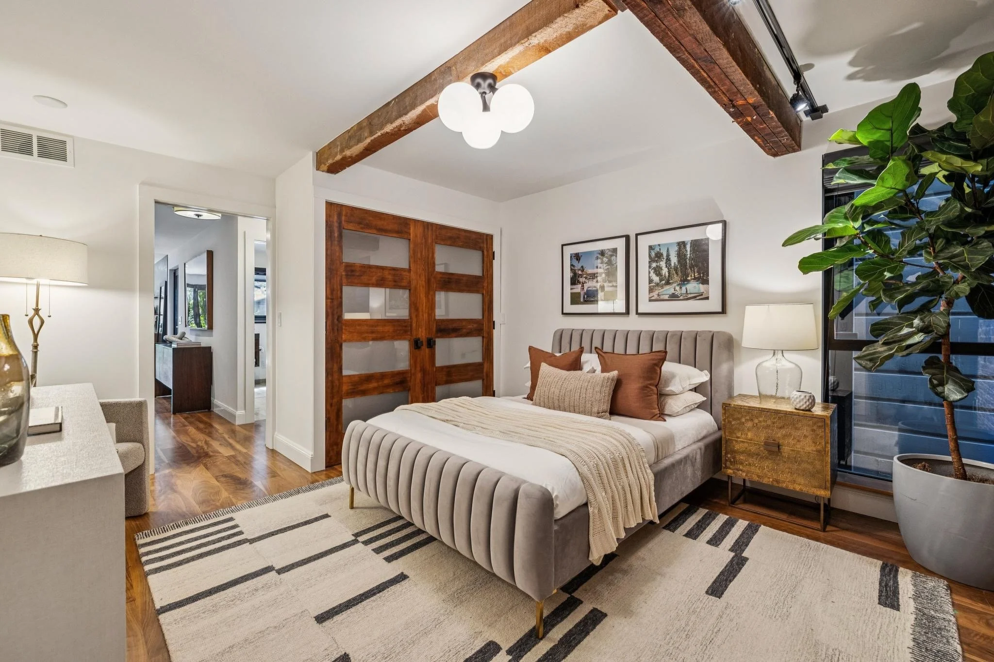Modern bedroom with a beige tufted bed, decorative pillows, a wooden side table, lush green plant, and framed artwork, located in a well-lit space with wooden floors and a striped area rug.