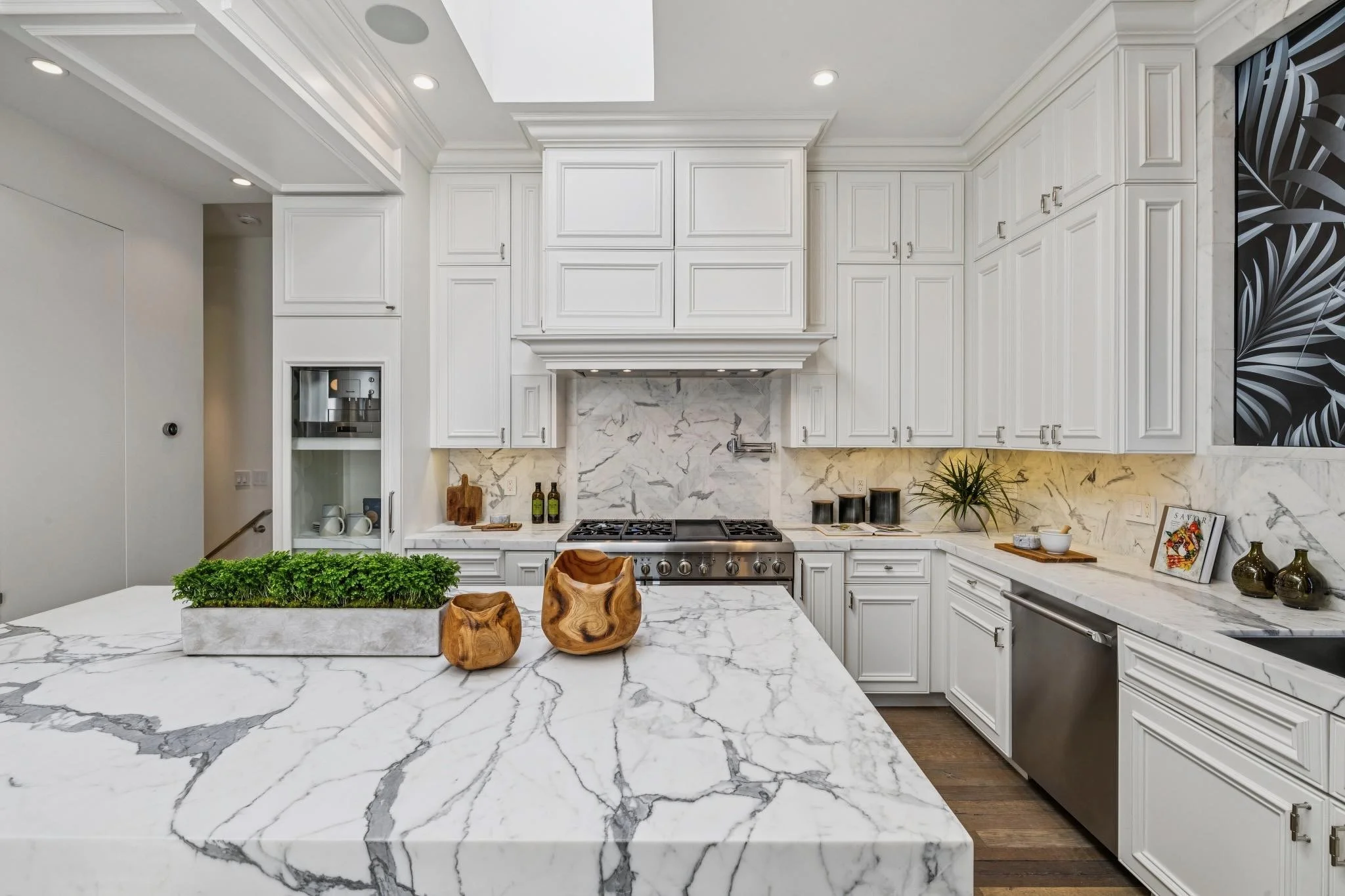 Modern kitchen with white cabinetry, marble countertops, and a matching marble backsplash. There is a stainless steel oven and some decorated items on the counter, including plants and framed pictures. The kitchen also features a kitchen island with 