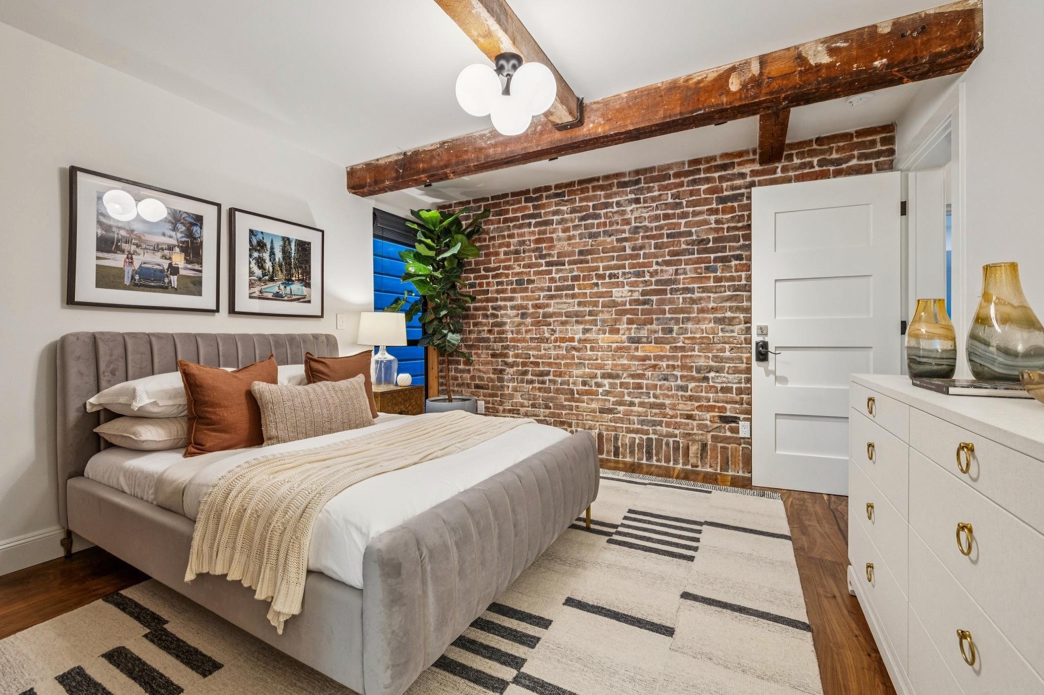 Modern bedroom with brick accent wall, beige upholstered bed with multiple pillows, side table with a lamp, framed photos on the wall, a large green plant next to the window, and a white dresser with decorative vases.