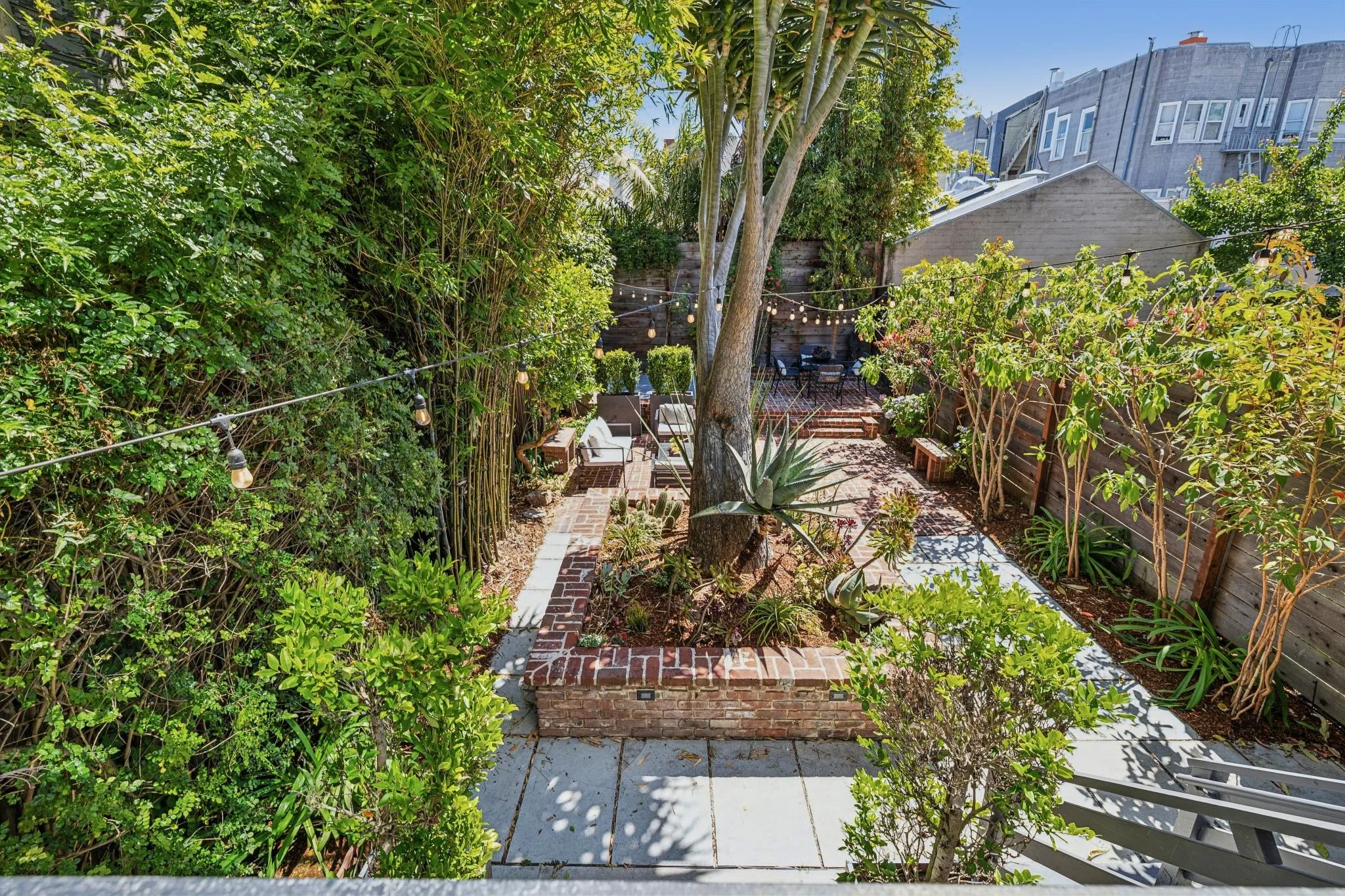 A lush backyard garden with a brick patio, surrounded by dense green foliage and trees, decorated with string lights, and featuring outdoor seating and plants.