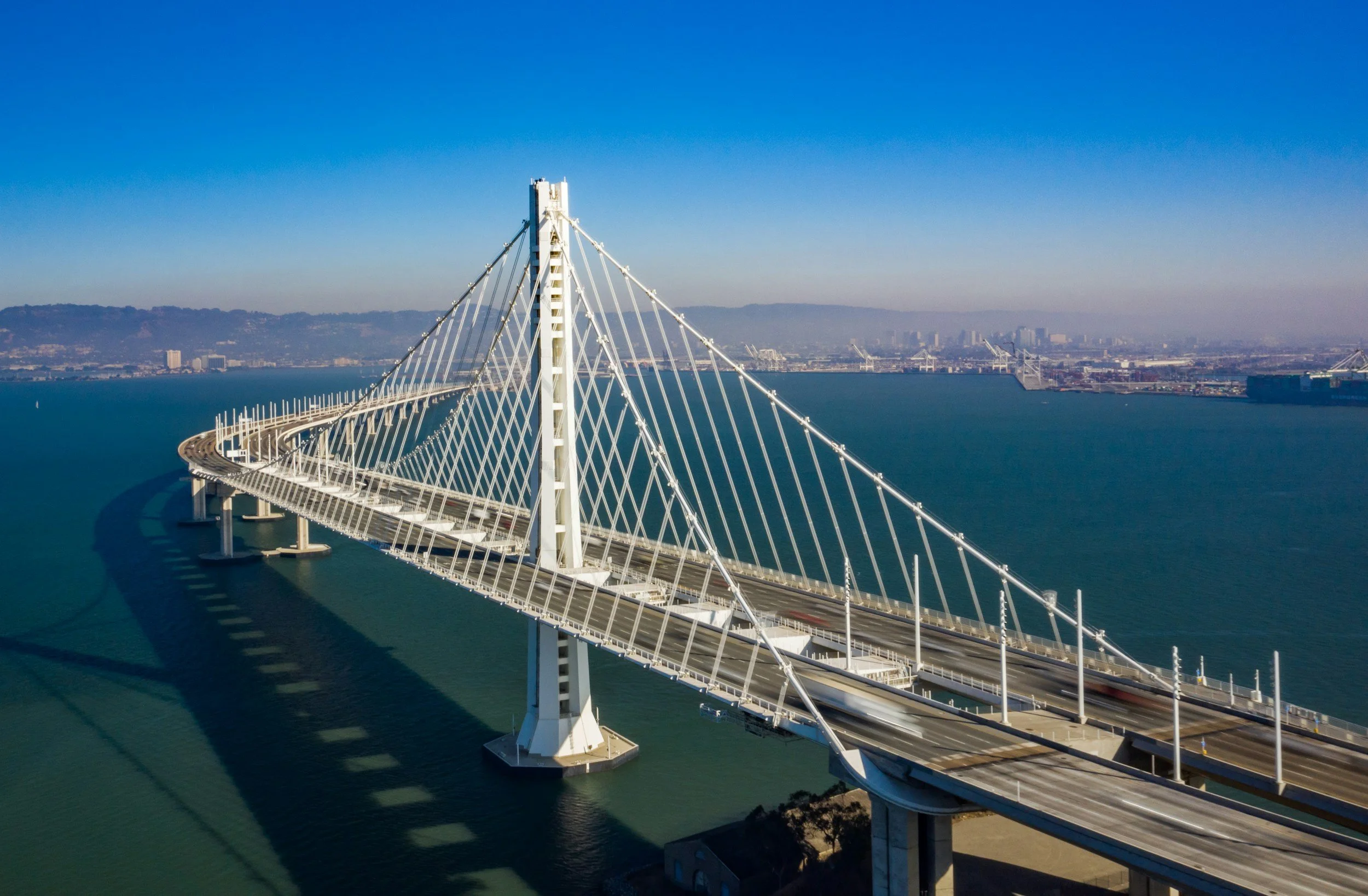 Large white cable-stayed bridge spanning over water with city skyline and hills in the background under a clear blue sky.