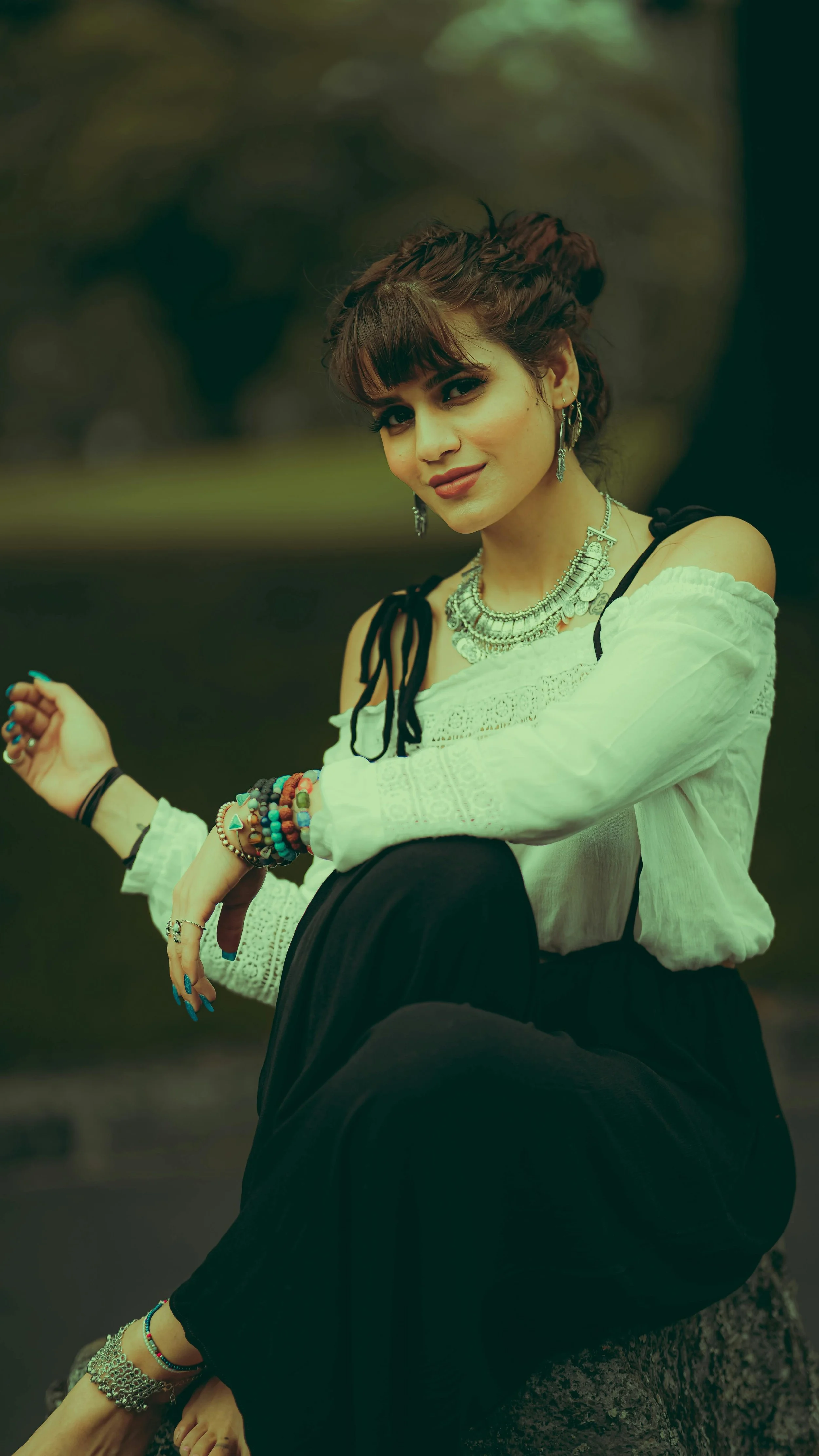 A young woman with short curly brown hair and bangs, wearing a white embroidered blouse with black straps, multiple colorful bracelets, and silver jewelry, sitting outdoors on a tree trunk.