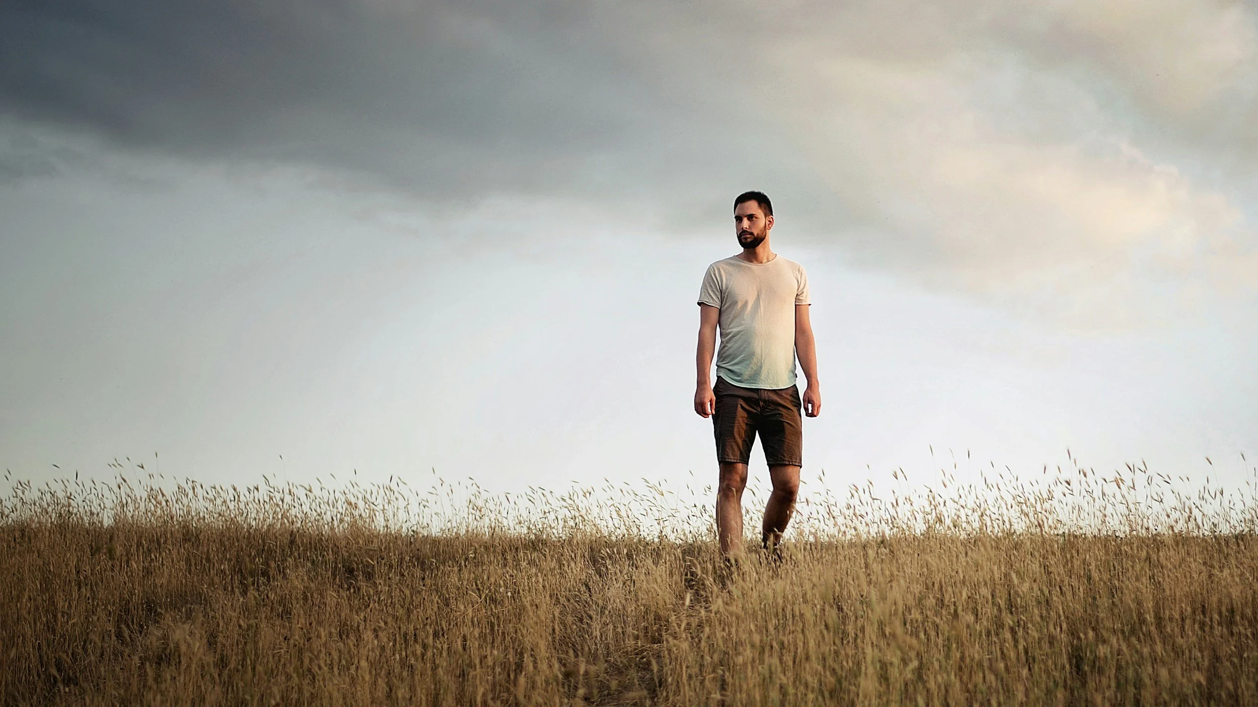 A man walking through a tall field of grass under a cloudy sky, dressed in a beige T-shirt and brown shorts.